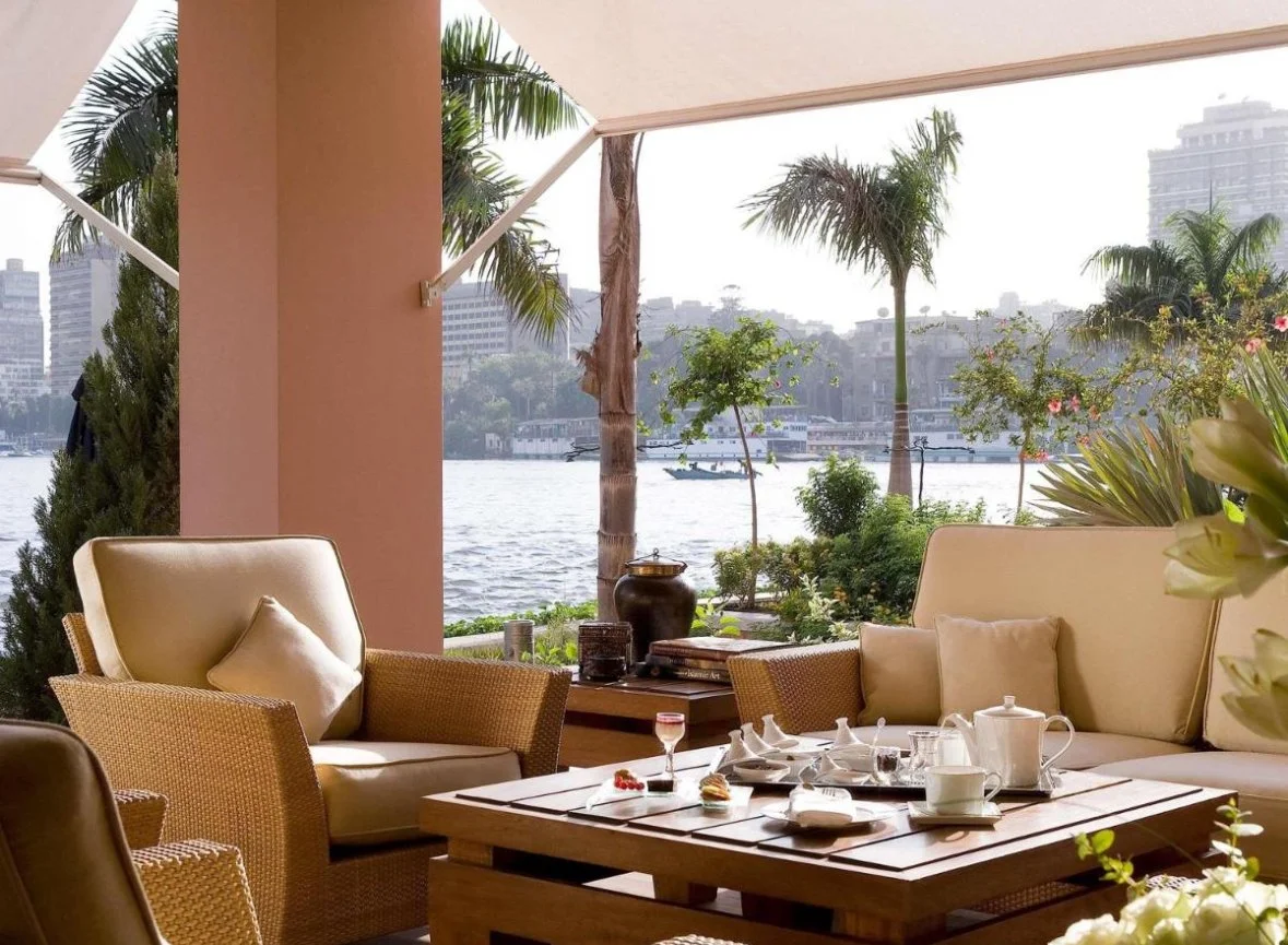Outdoor patio with a table set for tea, overlooking a river with trees and buildings in the background.