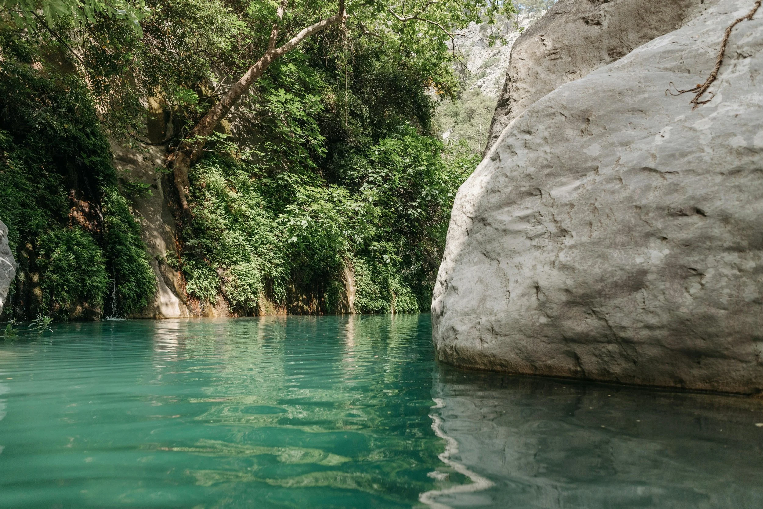 A calm river with turquoise water running between green trees and large gray rocks along the riverbank.