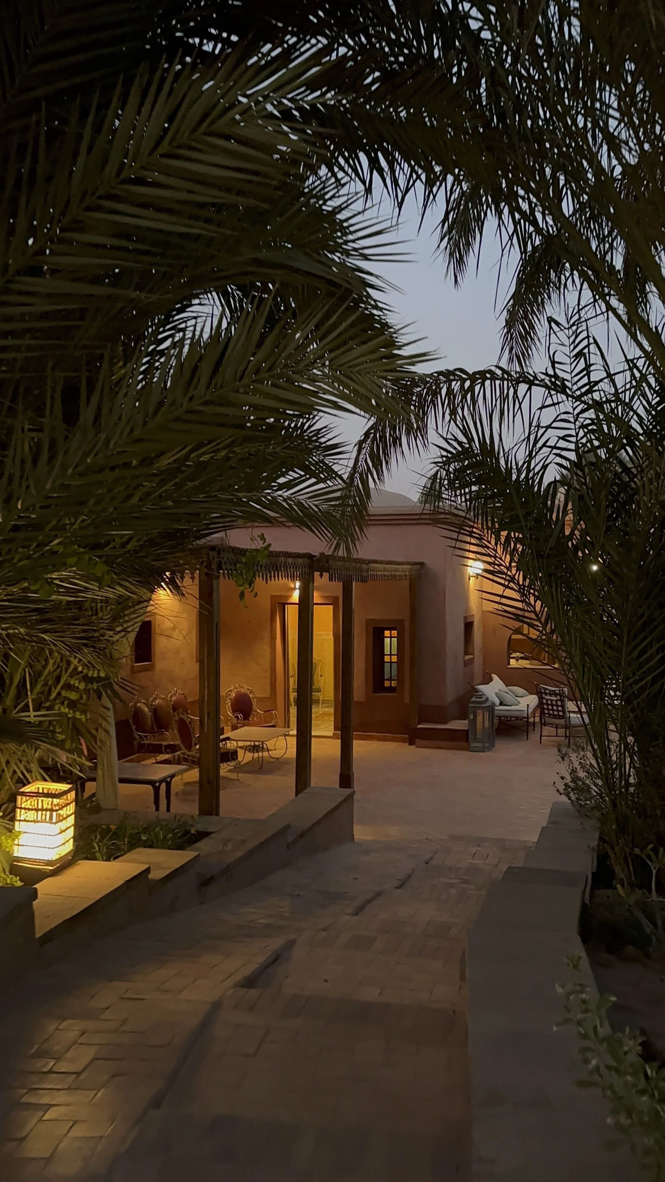 A cozy outdoor patio at dusk with furniture and decorative lighting, framed by palm leaves.