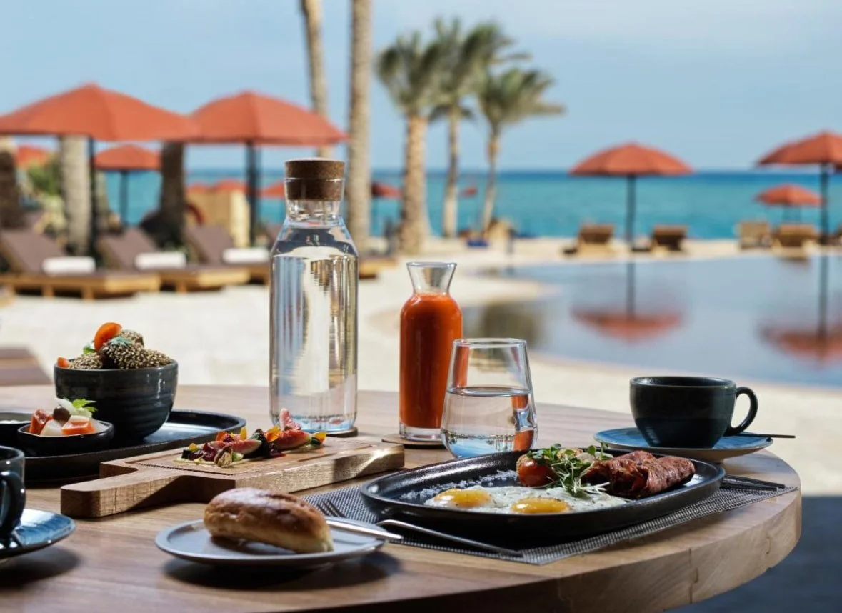 Breakfast table set with eggs, grilled meat, coffee, water, hot sauce, and salads, overlooking a pool and beach with umbrellas and chairs.
