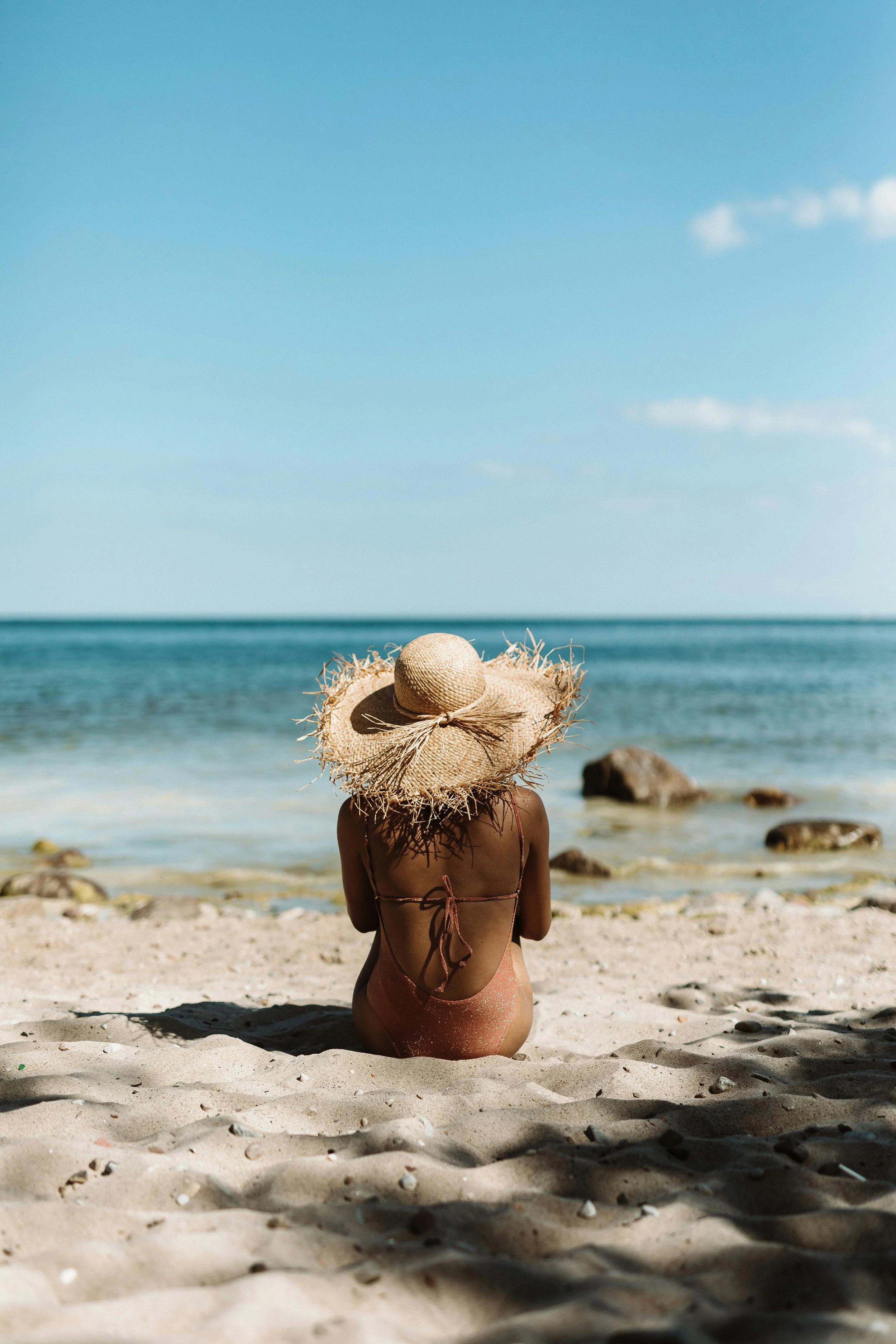 Woman in swimsuit with large straw hat sitting on sandy beach facing the ocean in Bali.