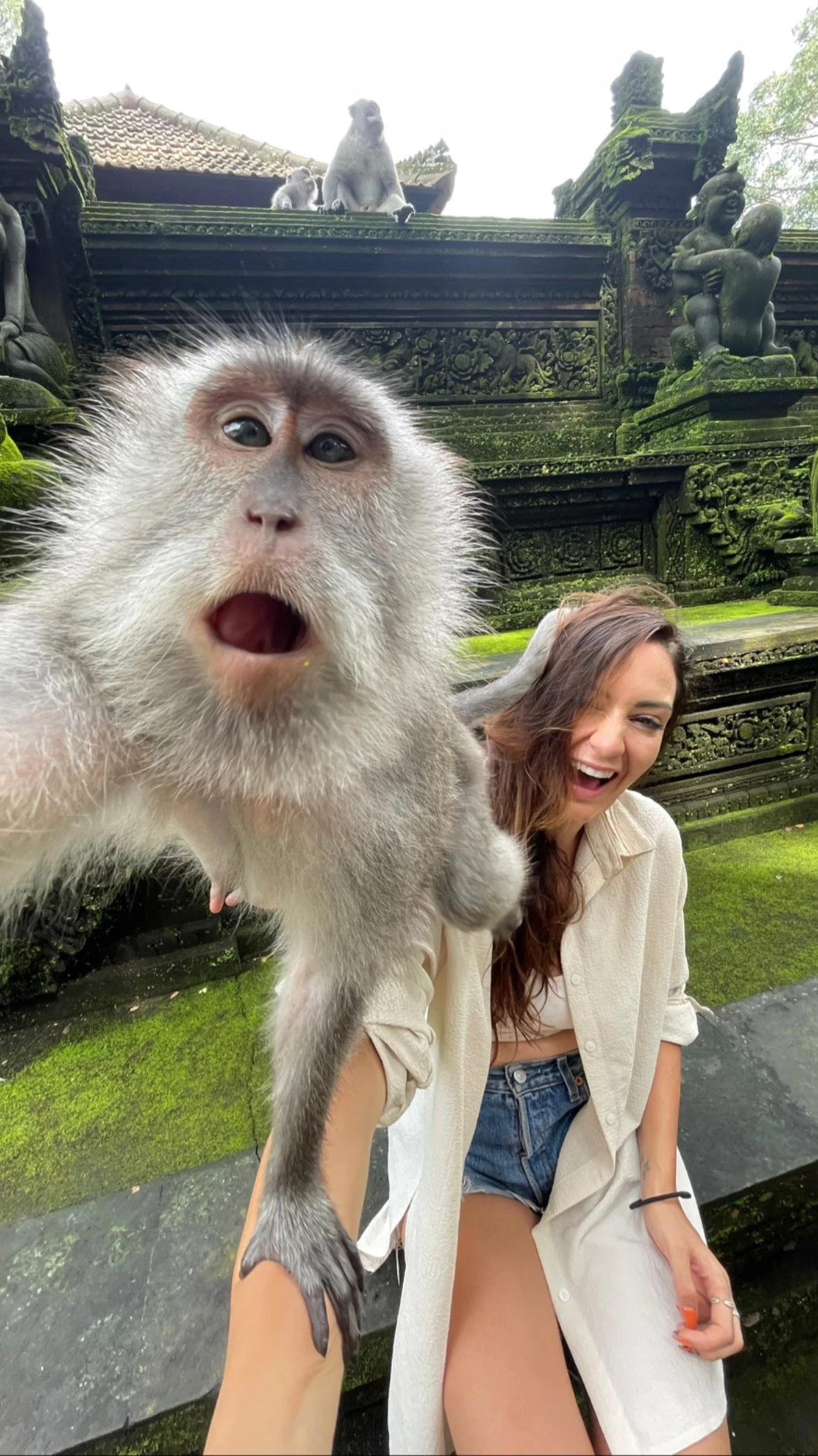 A woman taking a selfie with a monkey at the sacred monkey temple in Ubud with intricate stone carvings and statues, moss-covered stone structures, and greenery in the background.