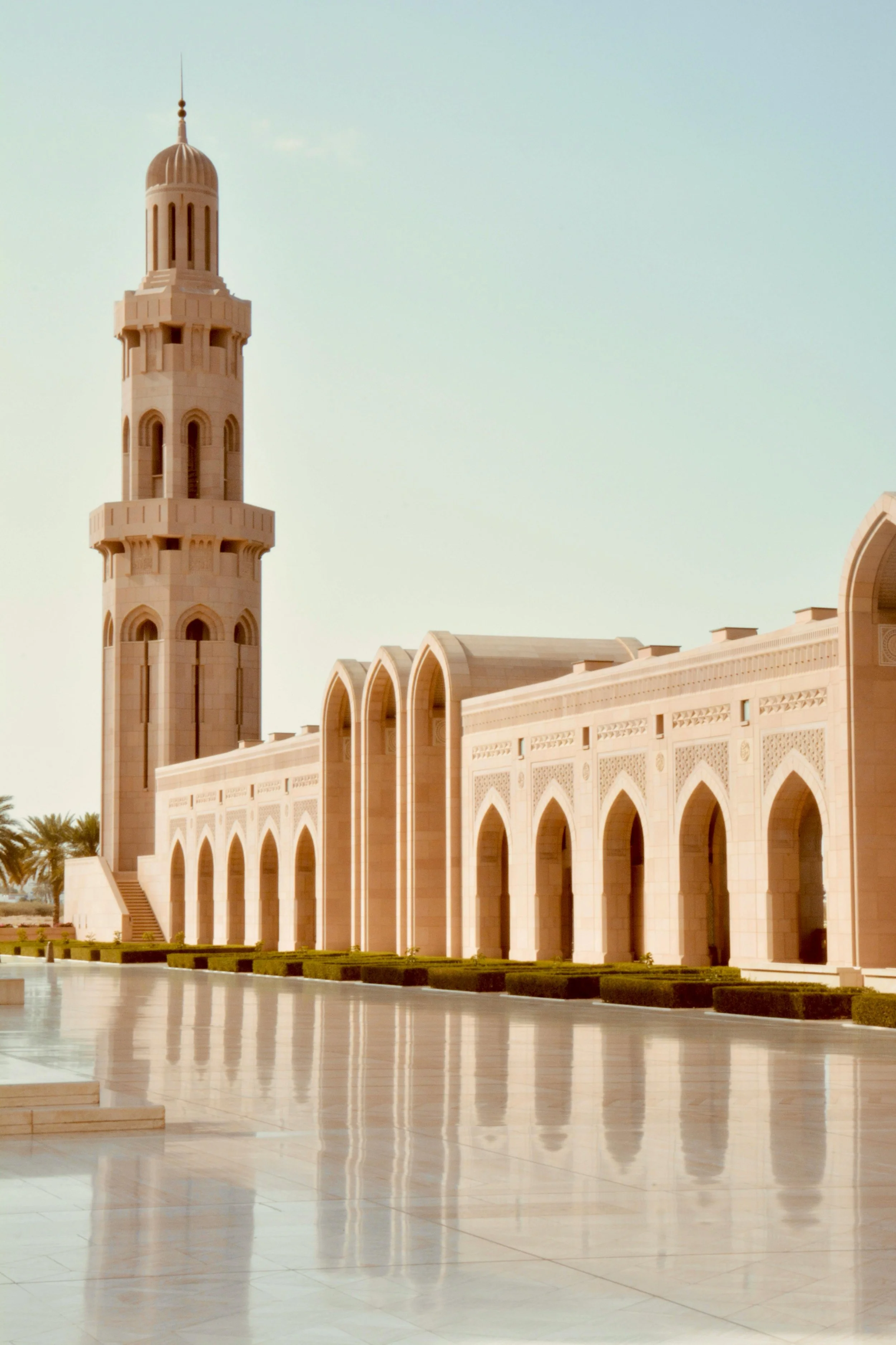 Minaret and mosque architecture with arches in Oman and reflection on polished floor, clear sky in background.