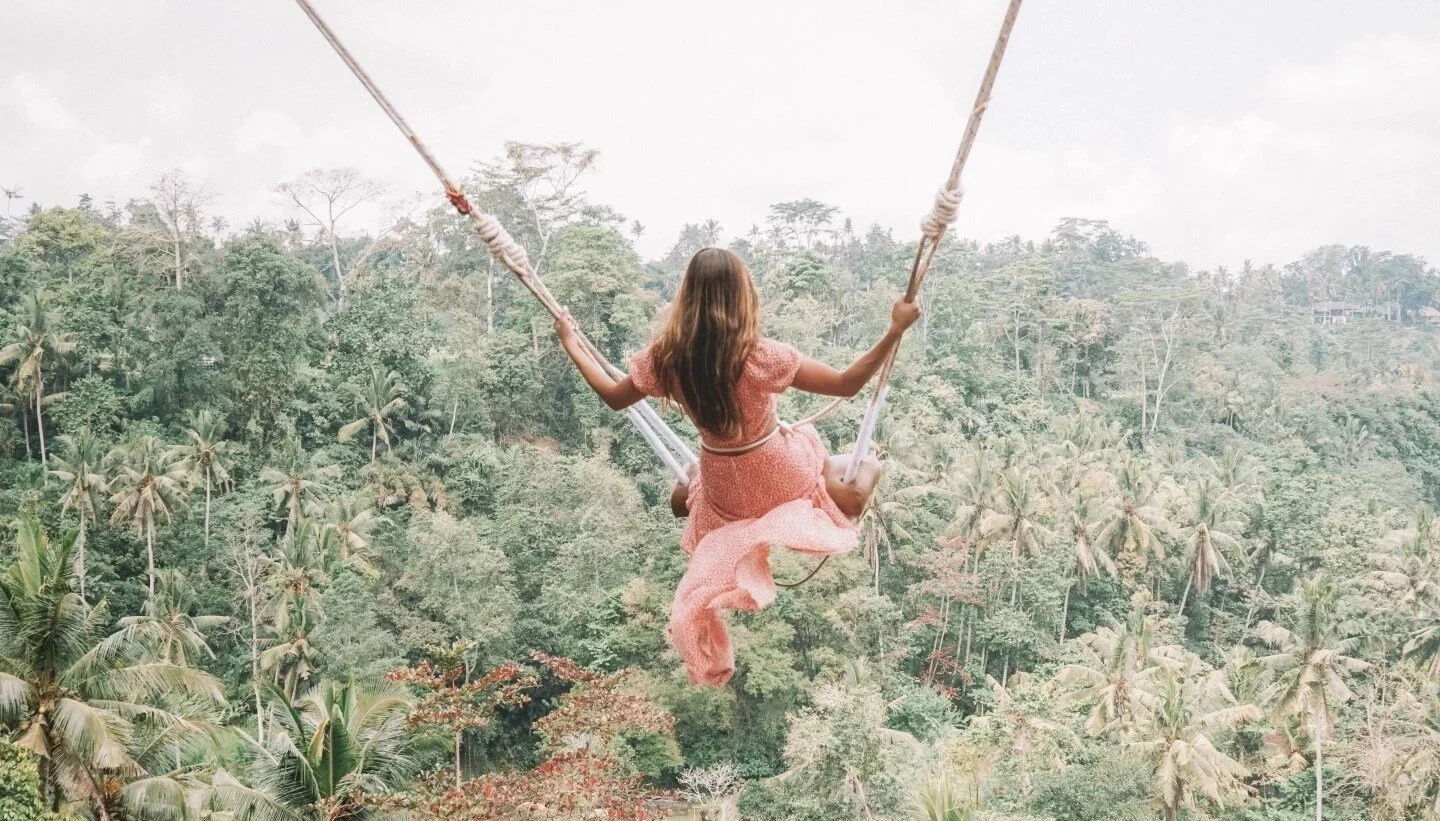 A girl in a pink dress swinging on the famous rice terrace in Bali over a lush green jungle.