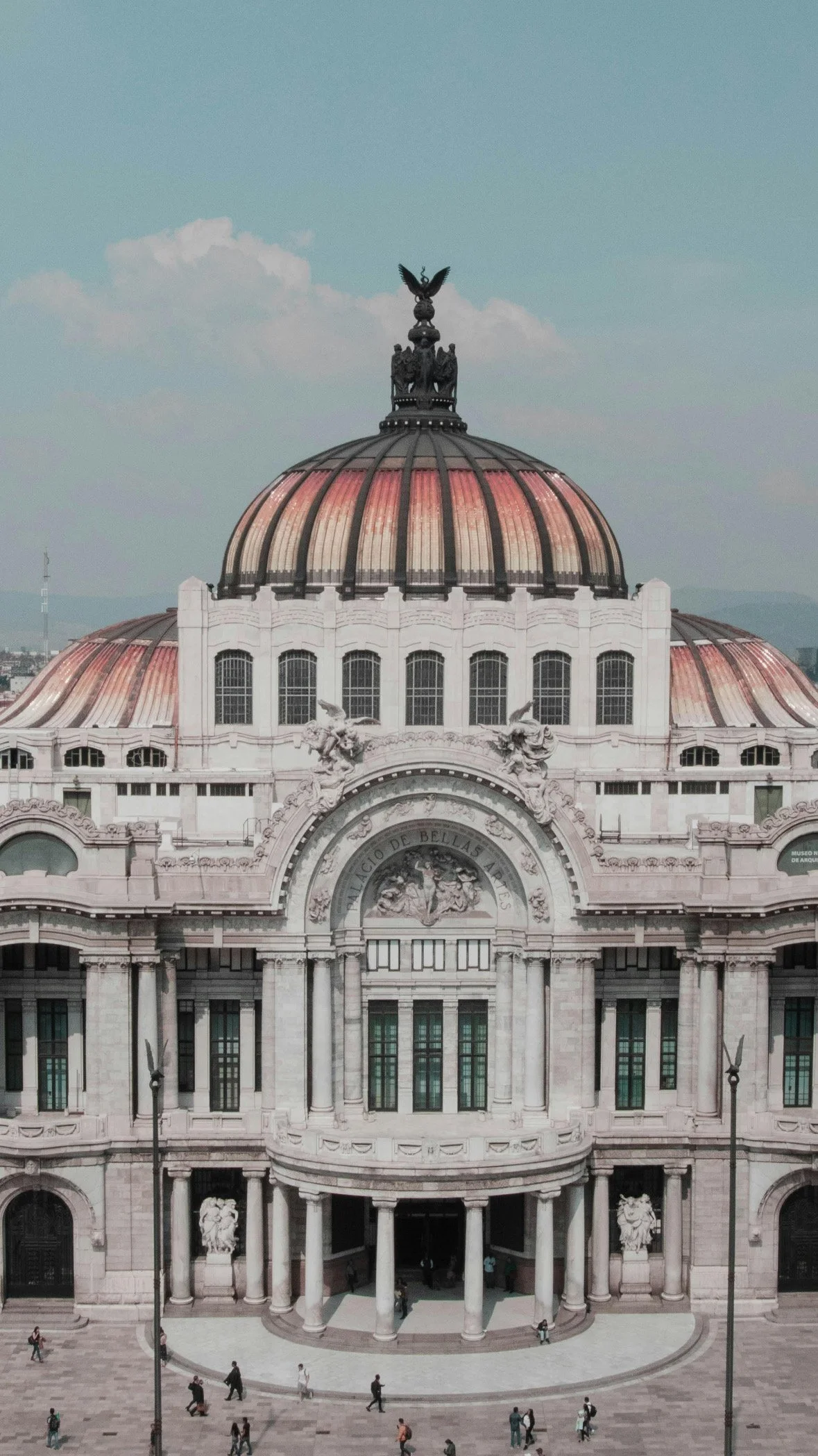 Historic building with a large dome, decorative statues, and pilars, with people walking in front in Oman. The building has the words 'Palacio de Bellas Artes' on it.