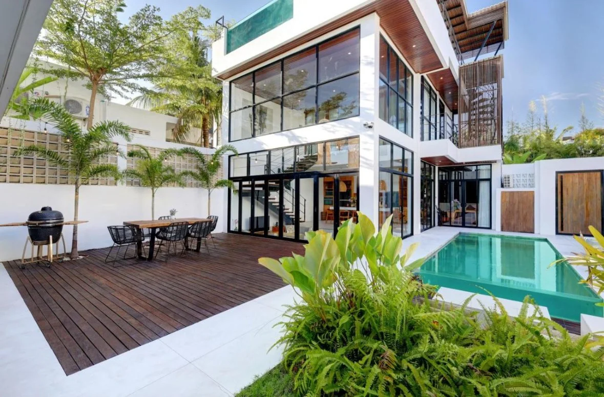 Modern two-story house with large glass windows, surrounded by lush greenery, featuring a swimming pool, outdoor dining area with table and chairs, and a wooden deck.