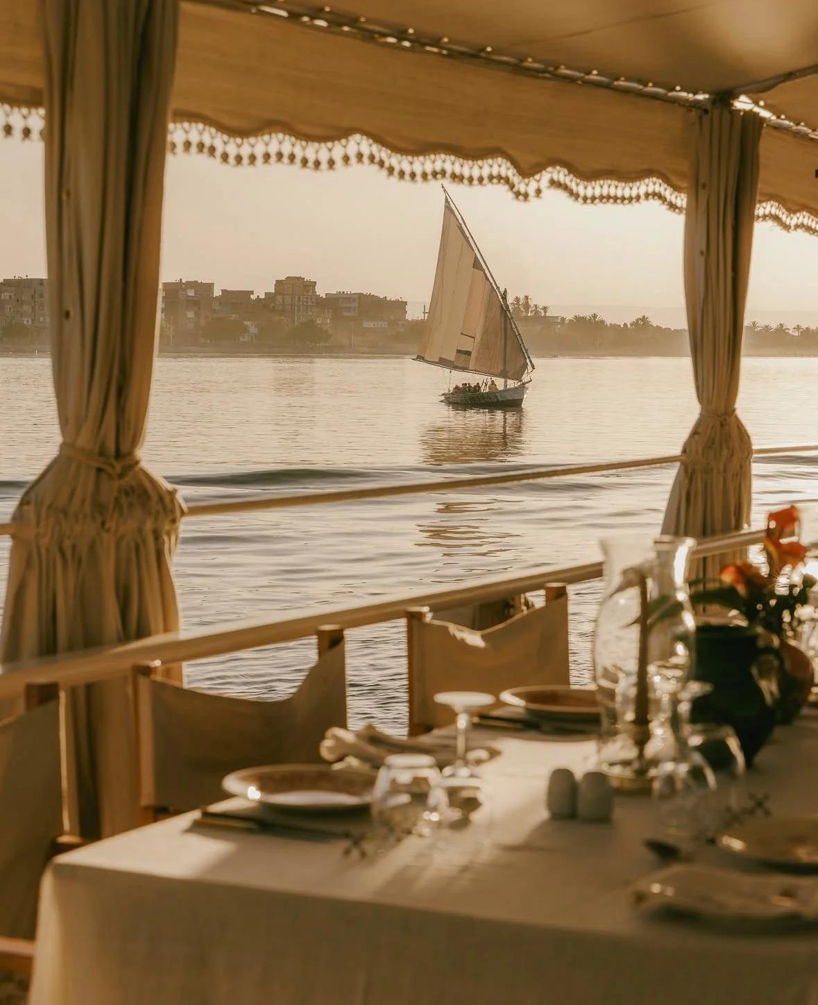 A table set for dining indoors with a view of a river, a sailboat sailing on the river, and a city skyline in the background during sunset.
