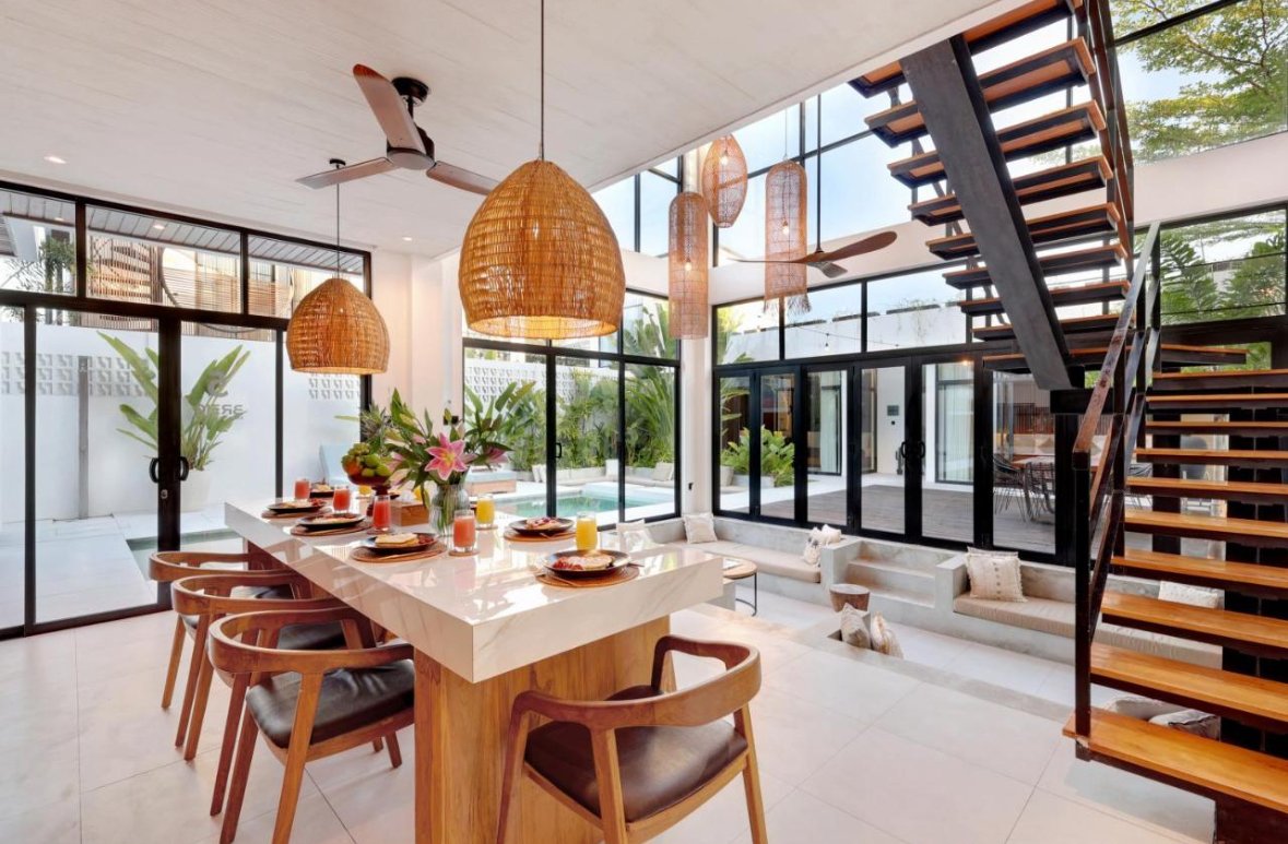 Modern dining area with a white marble table set for breakfast, surrounded by wooden chairs with leather seats. Large woven pendant lights hang above. Floor-to-ceiling glass windows and doors reveal a backyard with a pool and lush greenery. A stairca