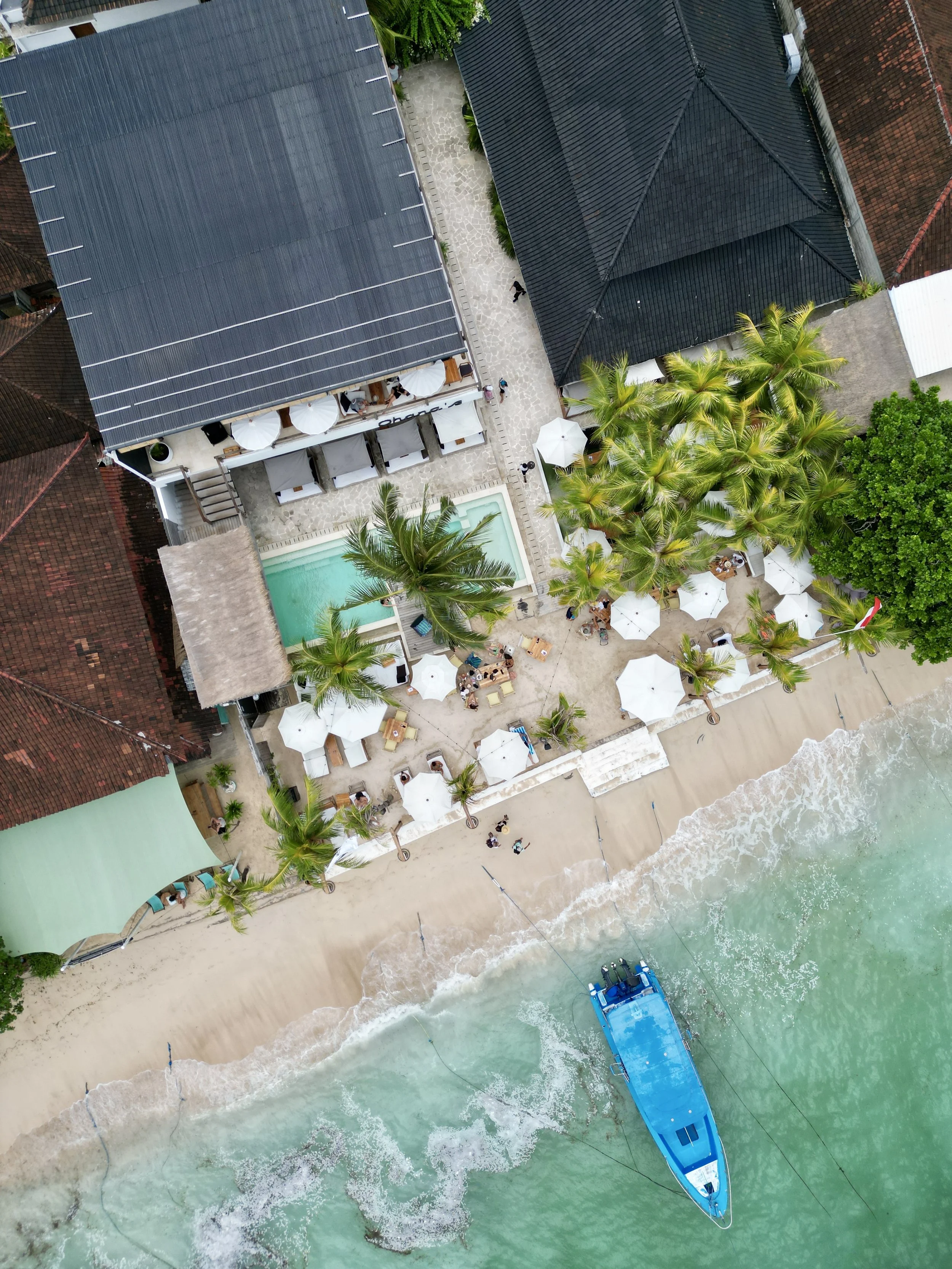An aerial view of a beachside resort in Bali with a sandy beach, turquoise water, palm trees, sun umbrellas, lounge chairs, and a boat docked near the shore.