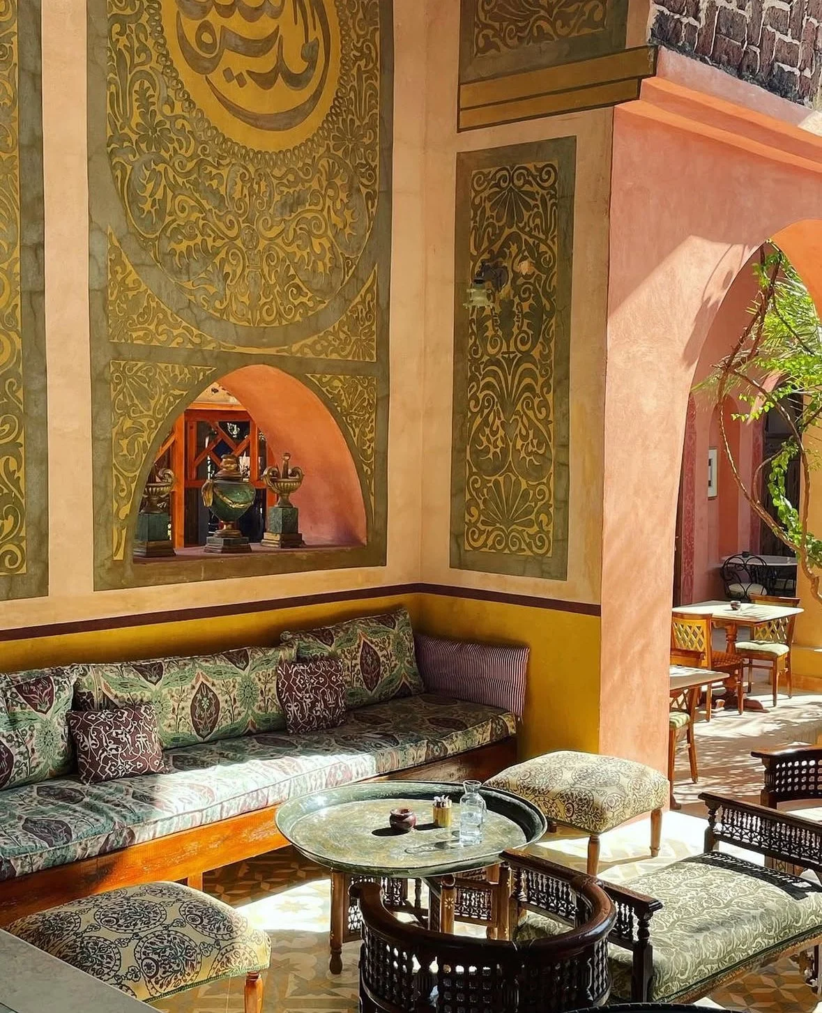 Interior of a Moroccan-style lounge with a decorative wall featuring ornate gold patterns, a long cushioned bench with patterned pillows, a round coffee table with items on it, and traditional furniture including chairs and ottomans, with sunlight streaming in.