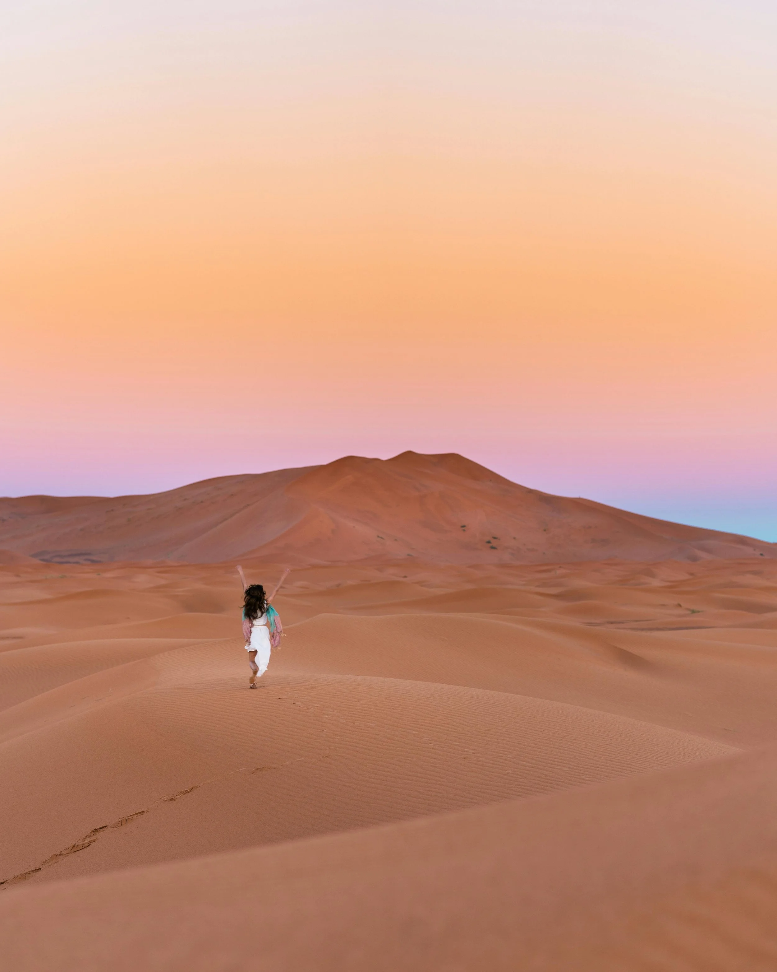 Person walking alone on sand dunes in Egypt during sunset or sunrise with a pastel-colored sky.
