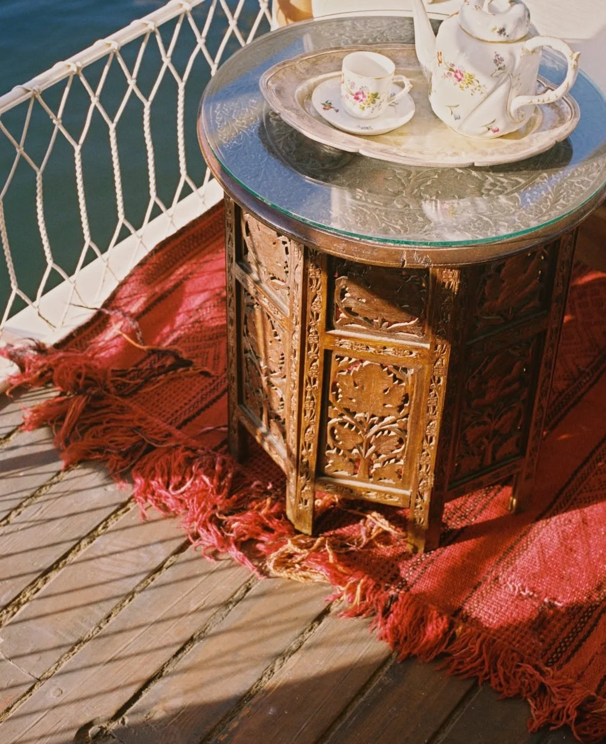 Tea set with floral teapot and matching cup and saucer on a silver tray, placed on an ornate wooden side table, with a red woven rug underneath on a wooden deck near water.