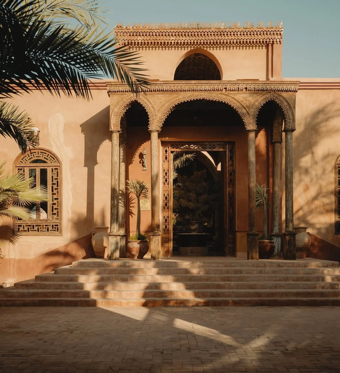 A building with traditional Middle Eastern architectural style, featuring arched doorways, decorative wooden accents, and potted plants at the entrance. There are steps leading up to the entrance, surrounded by palm trees and shadows cast by nearby foliage.