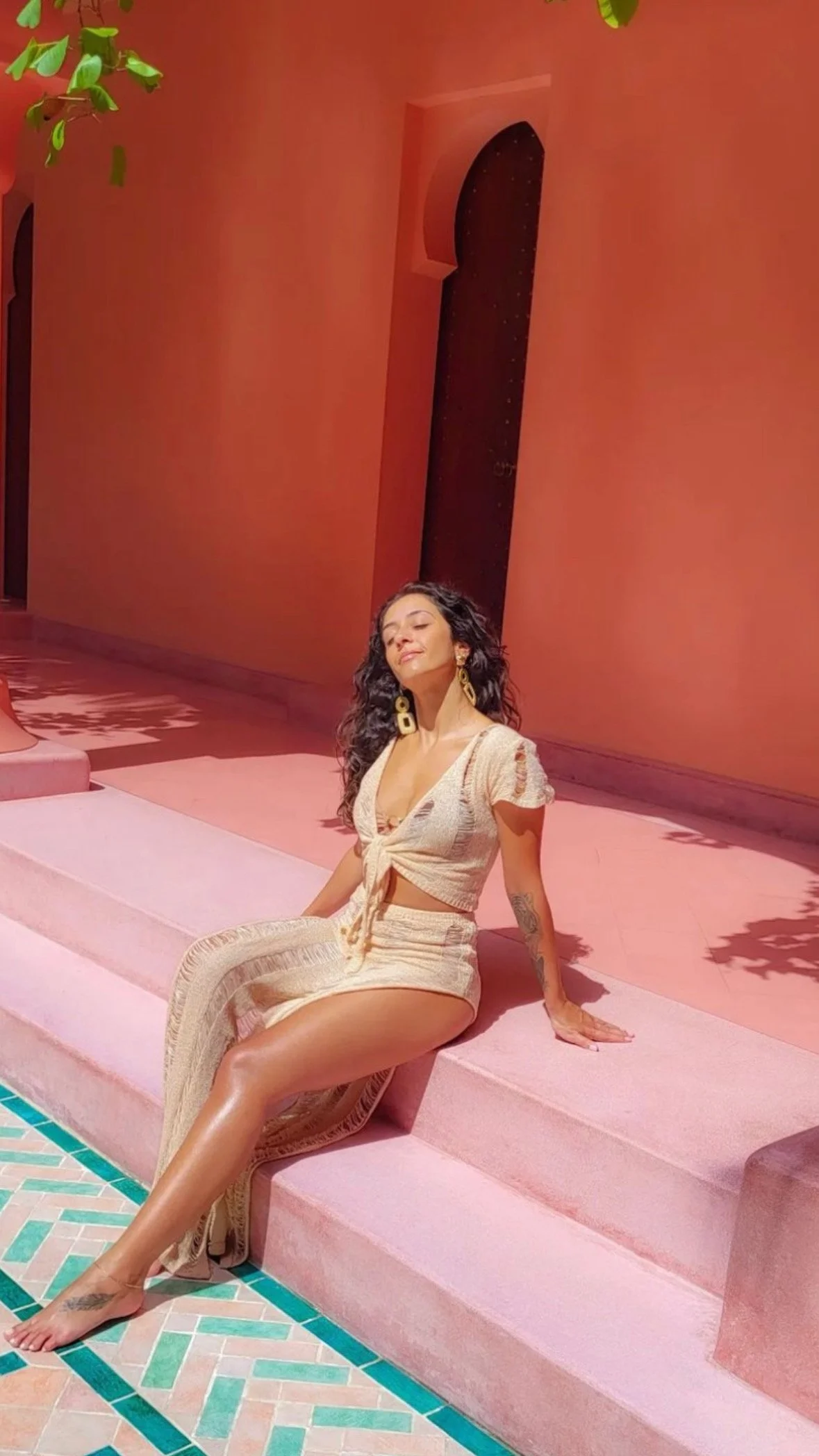 A woman with long curly dark hair wearing a beige crochet two-piece outfit with a cropped top and matching skirt, sitting on pink stairs near a pink wall with arched windows, in front of a tiled floor, smiling with her eyes closed and face tilted slightly upward
