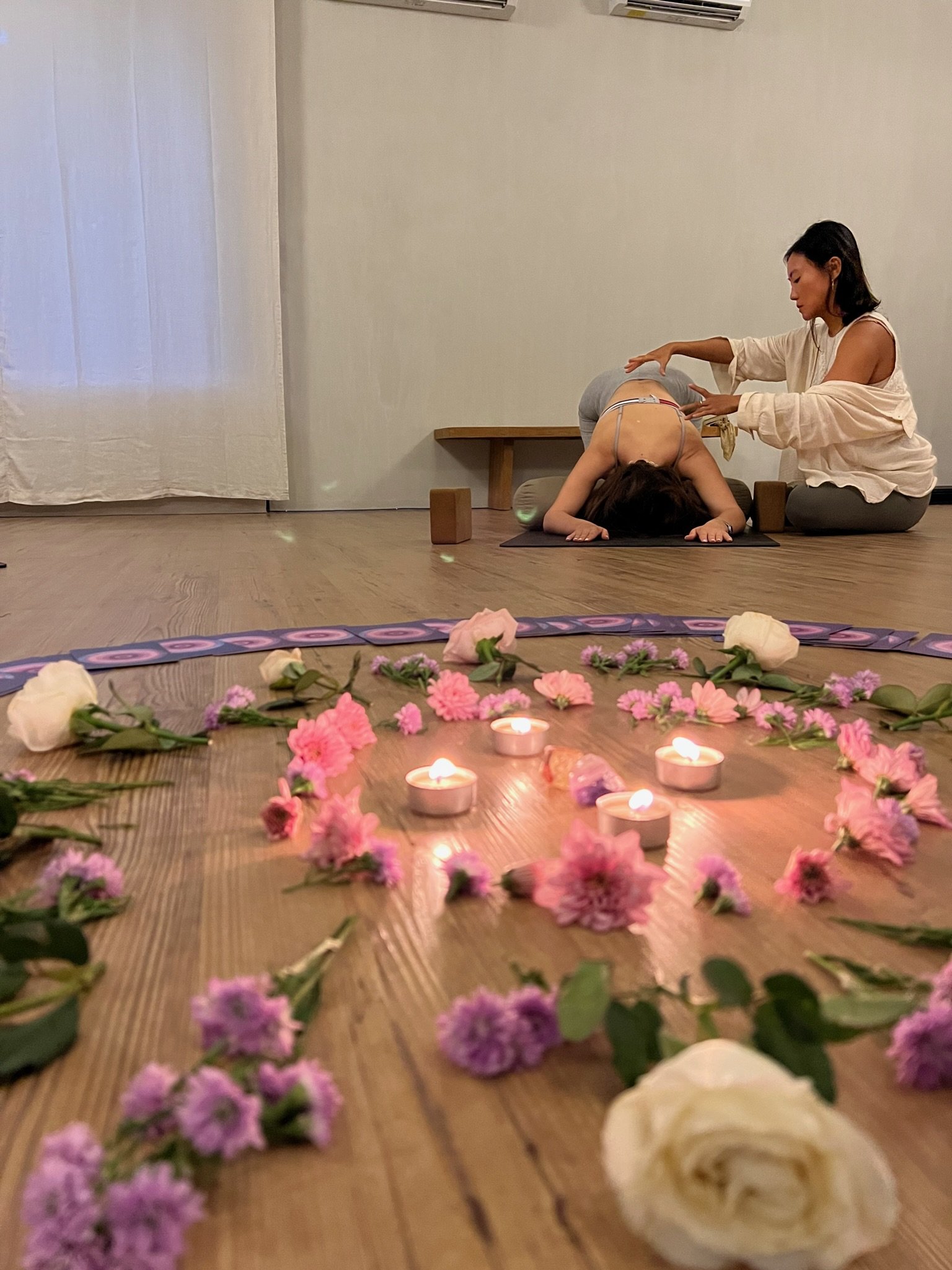 A woman receiving a yoga or meditation class with an instructor in a calm, decorated studio. The decor includes flowers and candles placed on the wooden floor in Canggu, Bali.