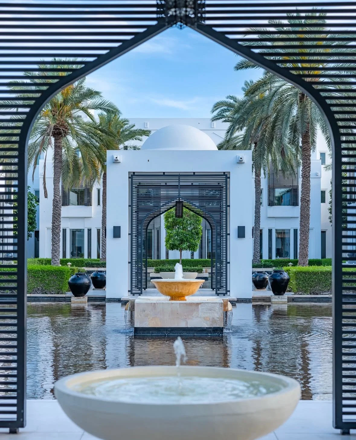 A view through a decorated archway shows a courtyard with a fountain, palm trees, and white buildings with modern architecture and greenery.
