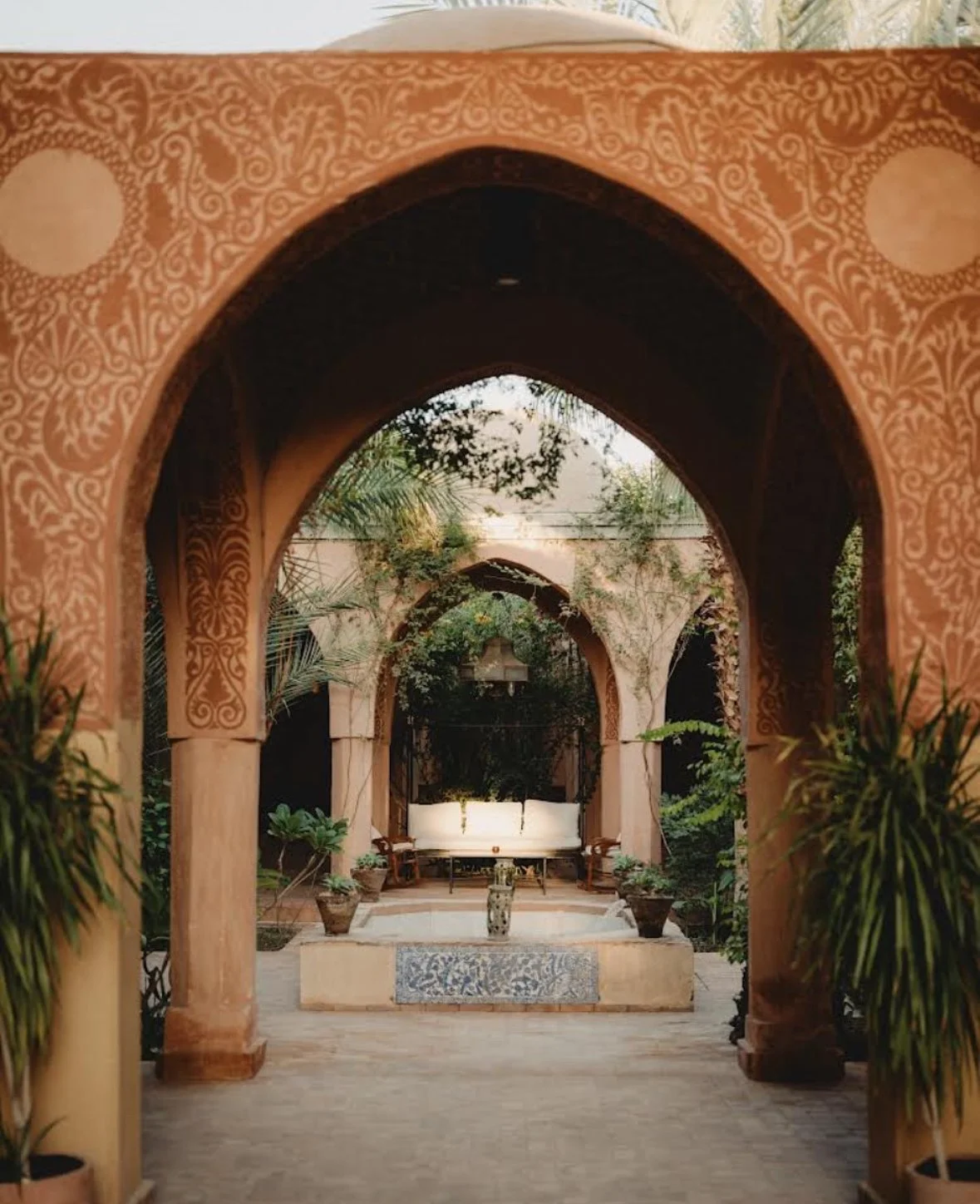 A courtyard with arches, potted plants, and a white fountain at the center, surrounded by lush greenery.