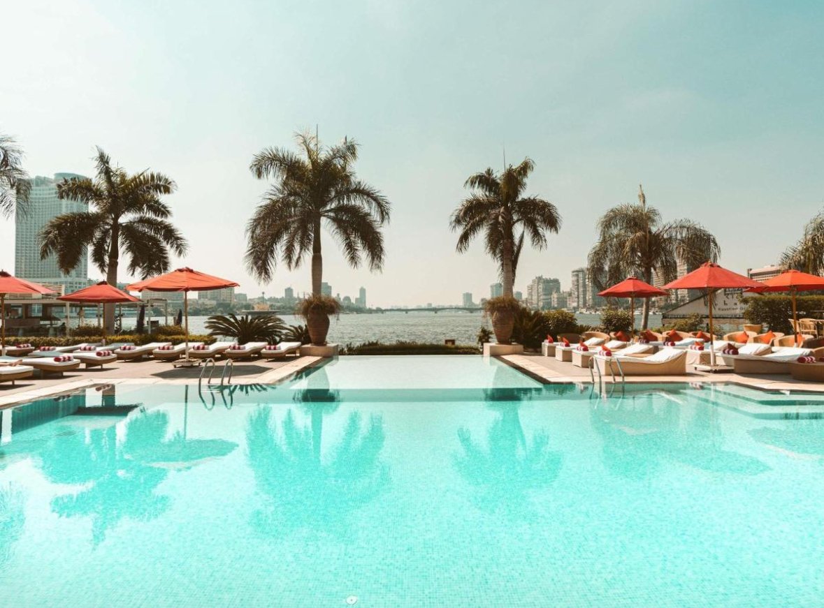 Swimming pool with lounge chairs and orange umbrellas overlooking a city skyline and water.