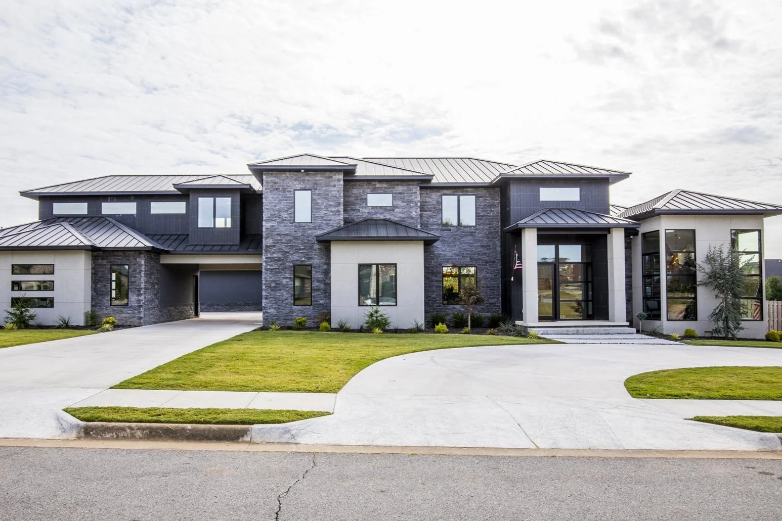 Modern two-story house with gray stone and white exterior, black metal roof, large windows, has a front lawn with a curved concrete driveway leading to a garage.