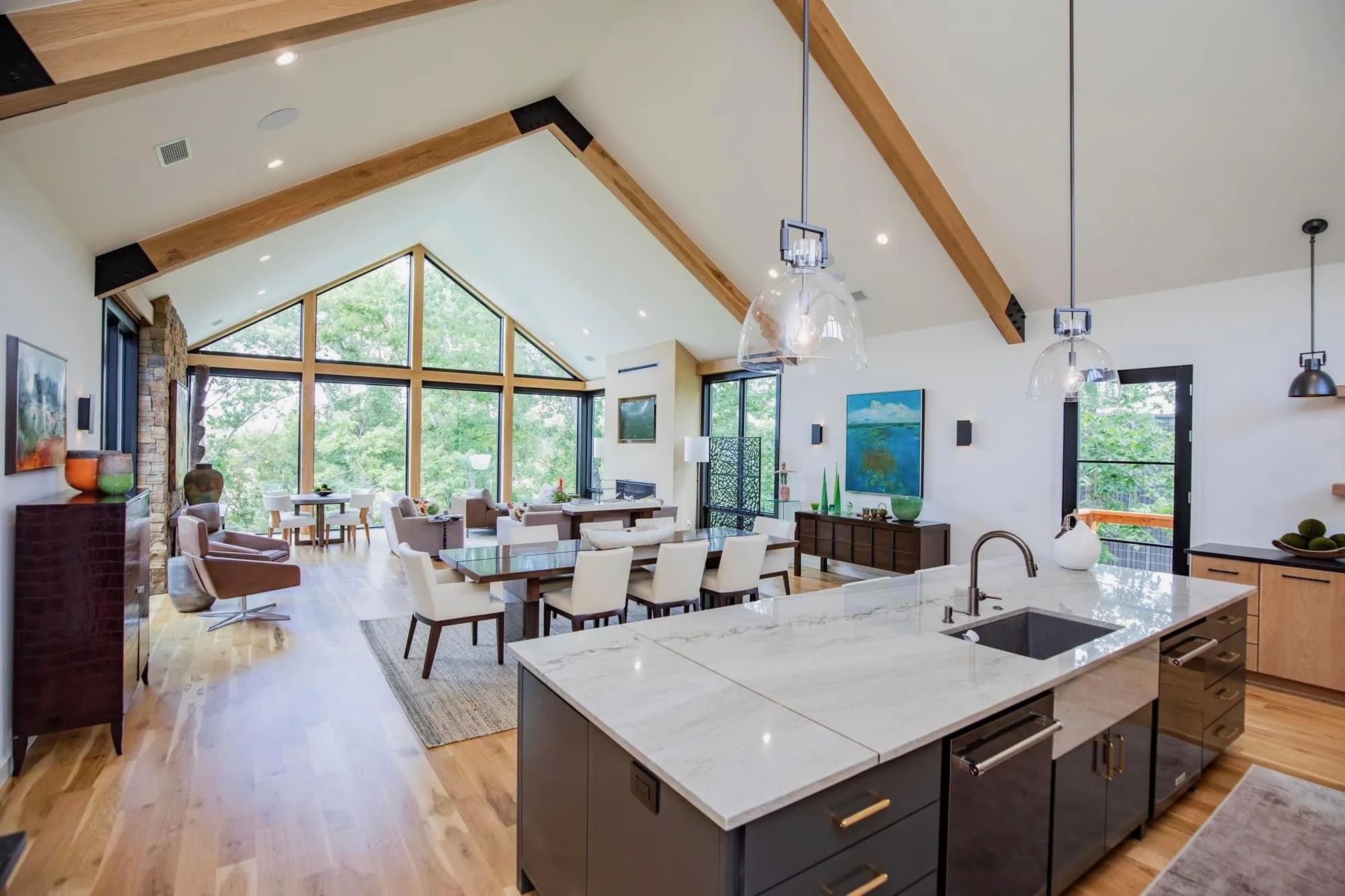Open-concept living room and kitchen with large vaulted ceiling, wood beams, floor-to-ceiling windows, and hardwood floors.