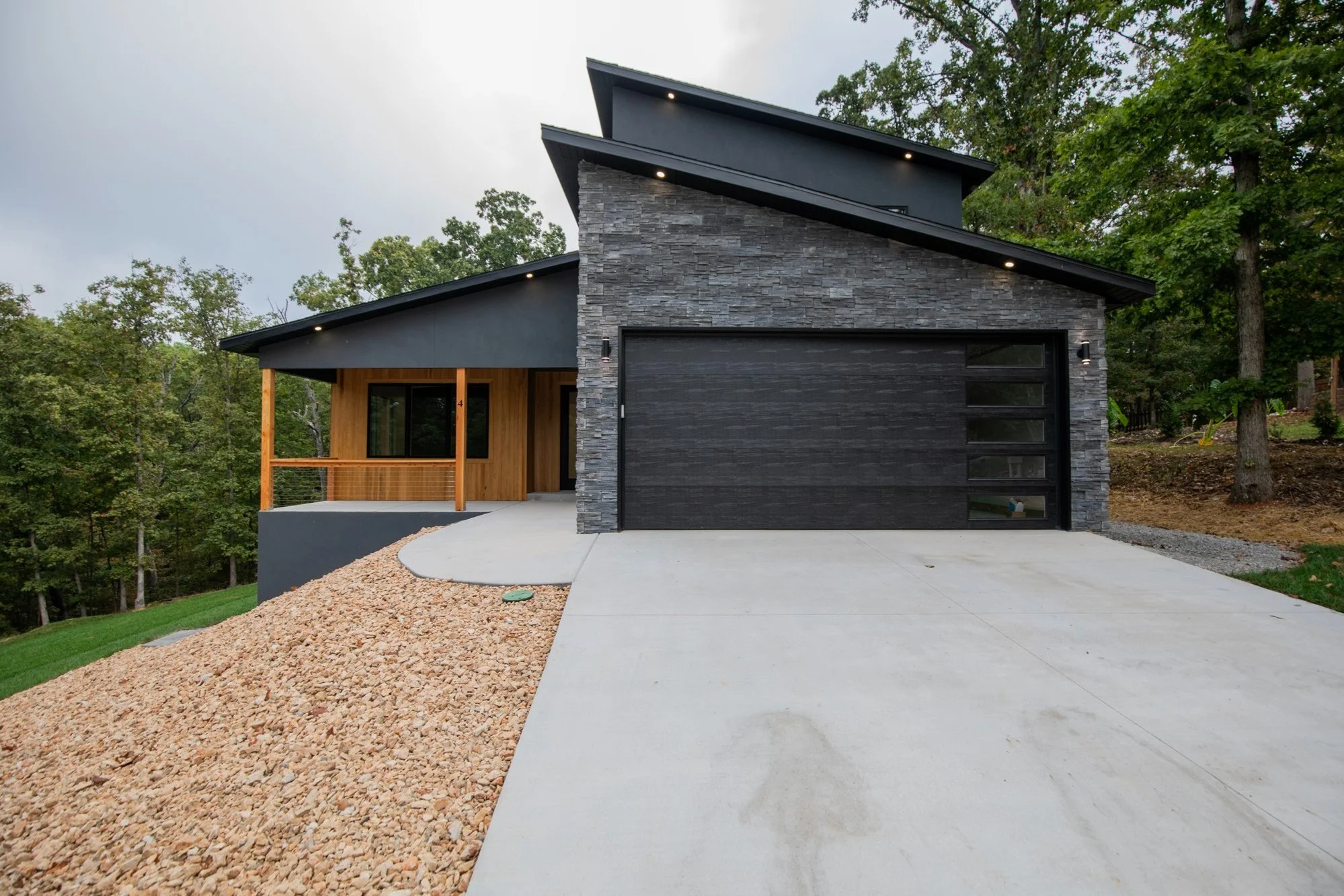Modern house with black garage door, stone accents, wooden porch, and sloped roof, surrounded by trees and a concrete driveway.