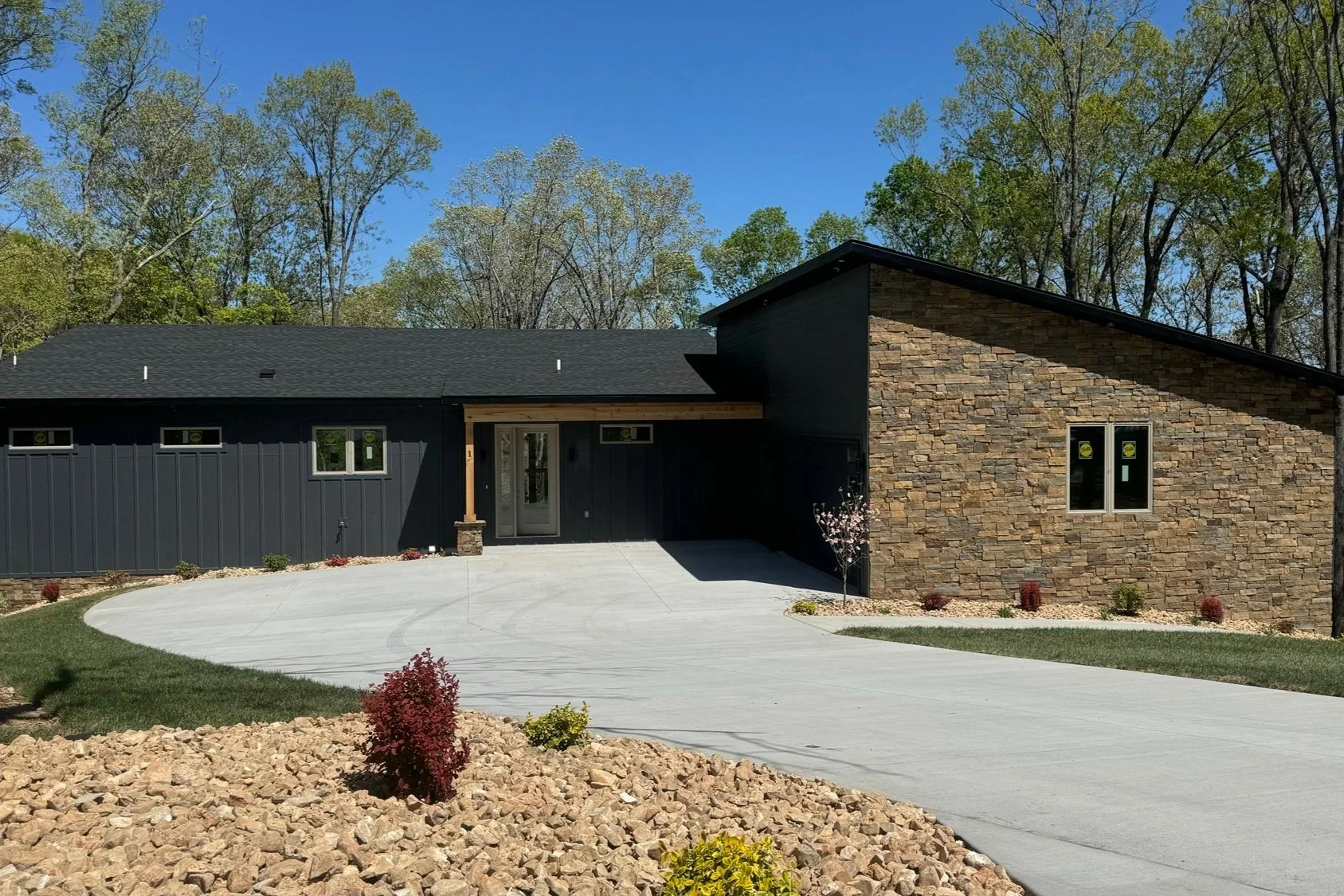 Modern house with black siding and a stone facade, concrete driveway, landscaped yard with rocks, small trees, and shrubs, surrounded by trees with a clear blue sky.