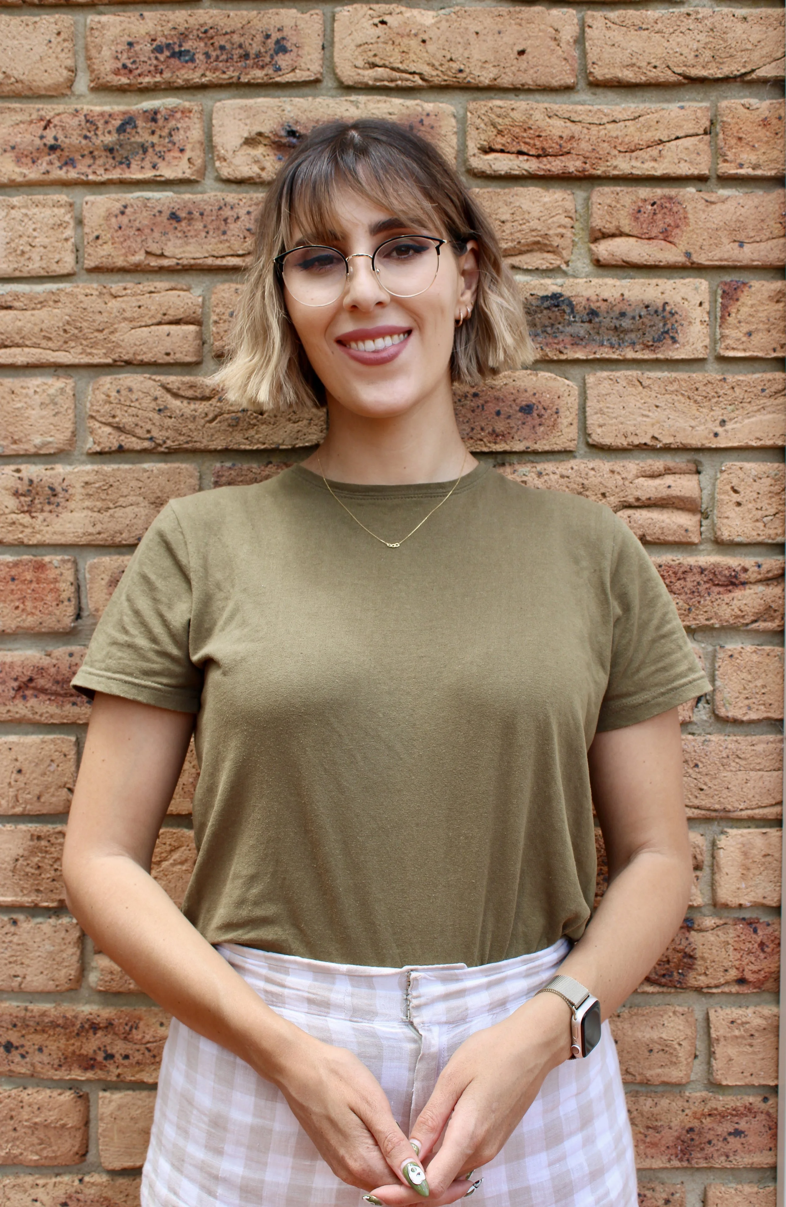 A photo of Kassandra standing against a brick wall. She has brown to blonde short ombre hair, has a fringe, wears black and gold glasses and is wearing a olive green t-shirt.