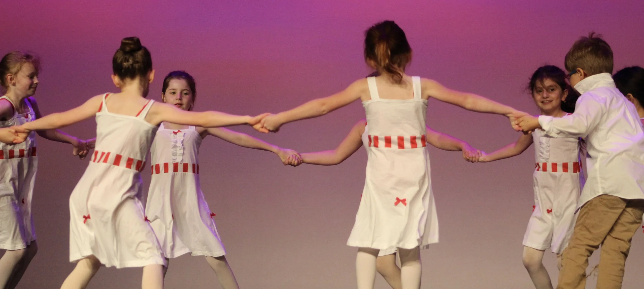 Children in white costumes with red stripes hold hands and dance on stage with purple lighting.