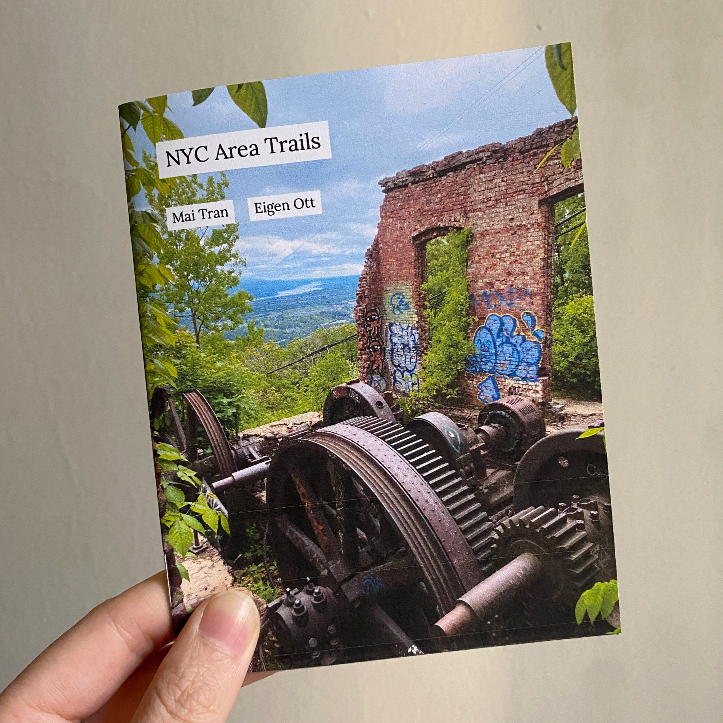 Cover of NYC Area Trails by Mai Tran and Eigen Ott, being held against a beige background. Cover is photo of ruins on Mt. Beacon, with a view of the Hudson Valley in the background.