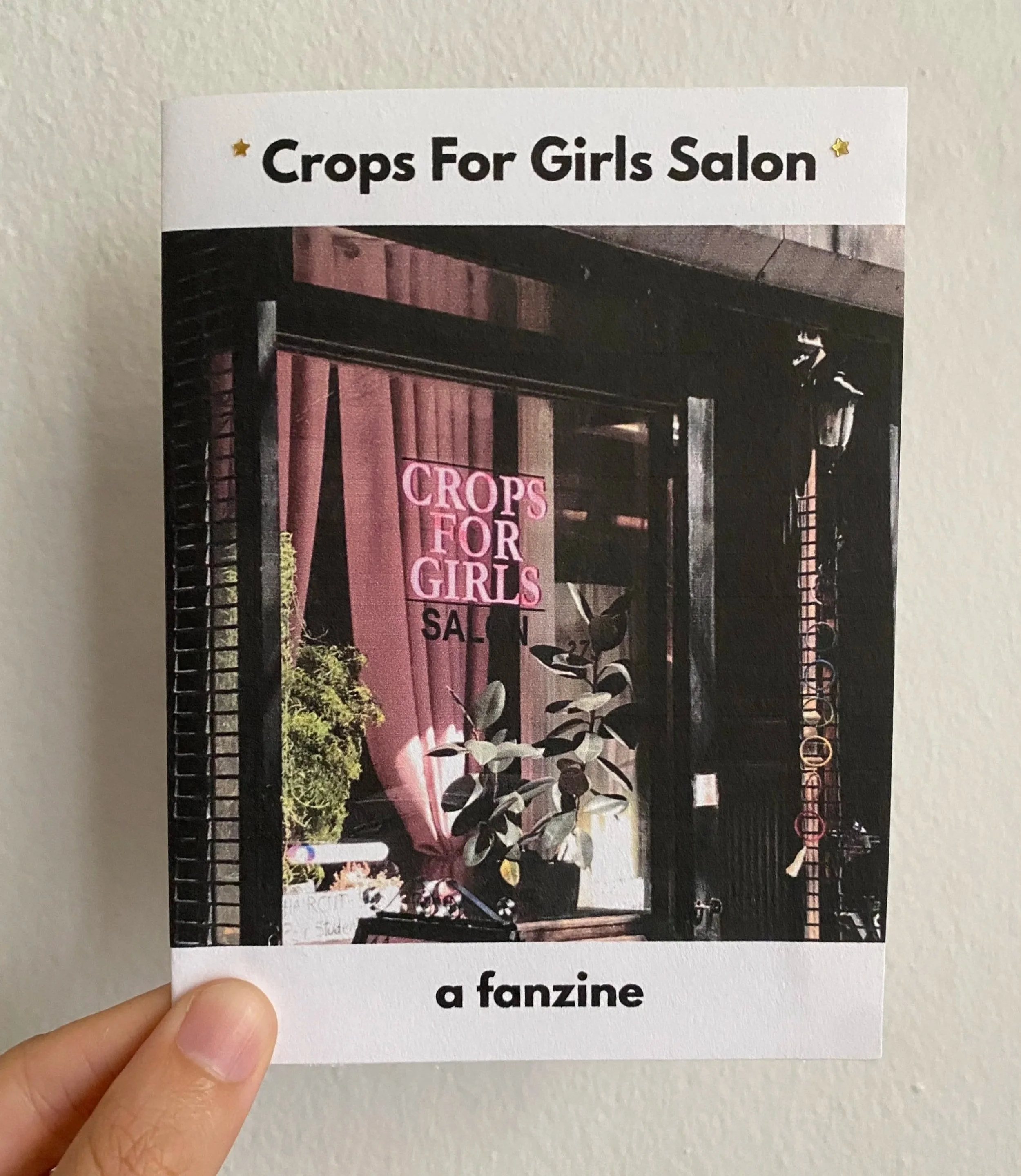 The Crops for Girls fanzine being held against a white wall. The cover has an image of the salon's storefront, which is dark wood with a pink curtain and plants in the window.