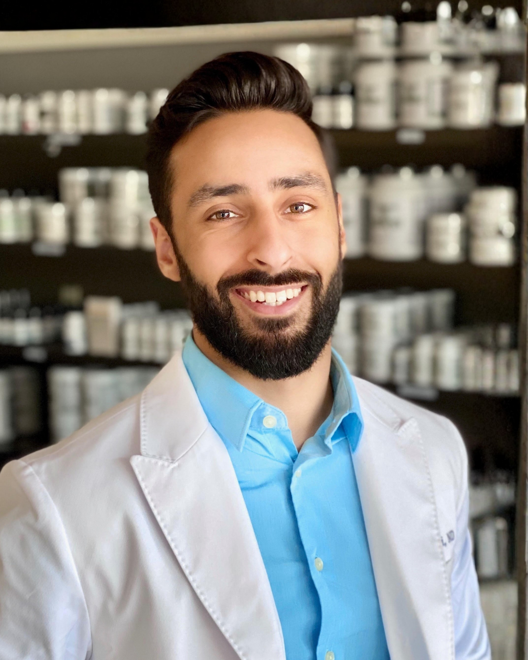 Portrait of a smiling man with a beard and dark, styled hair, wearing a patterned button-up shirt, standing outdoors with green foliage in the background.