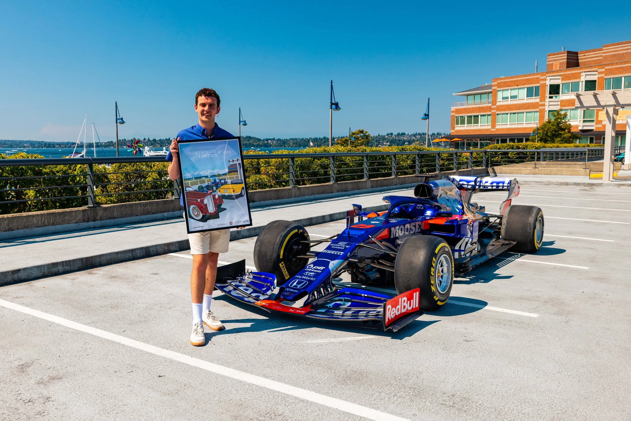 A young man wearing a blue polo shirt and shorts holding a framed poster of a vintage car next to a modern Red Bull racing car parked in a lot with a waterfront and a modern brick building in the background.