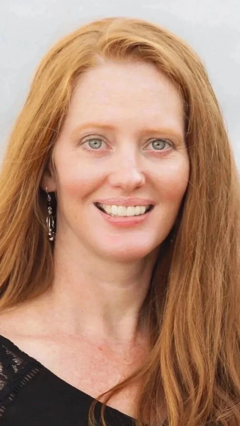 Close-up of a woman with long red hair, light skin, wearing earrings and a black top, smiling against a neutral background.