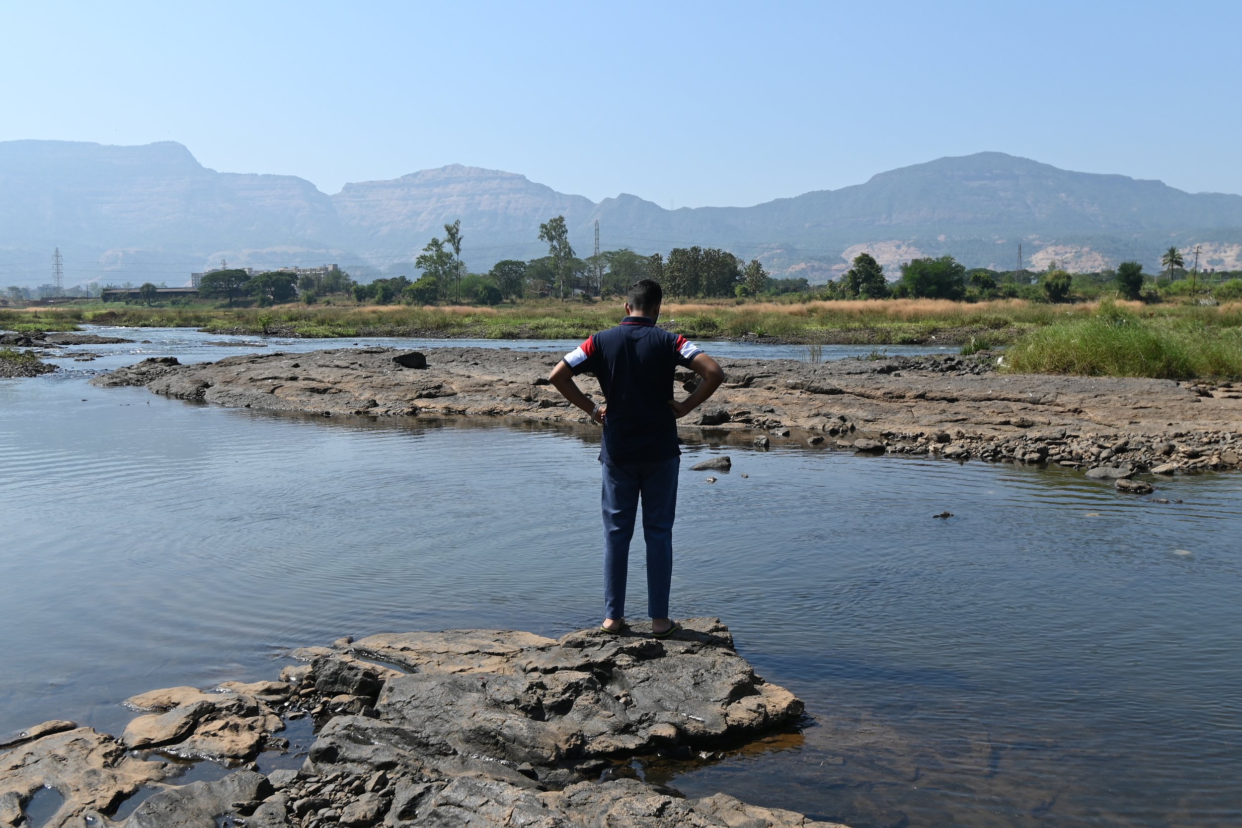 A man stands with back to the camera and his hands on his hips looking out at a landscape with a brook and hills in the background
