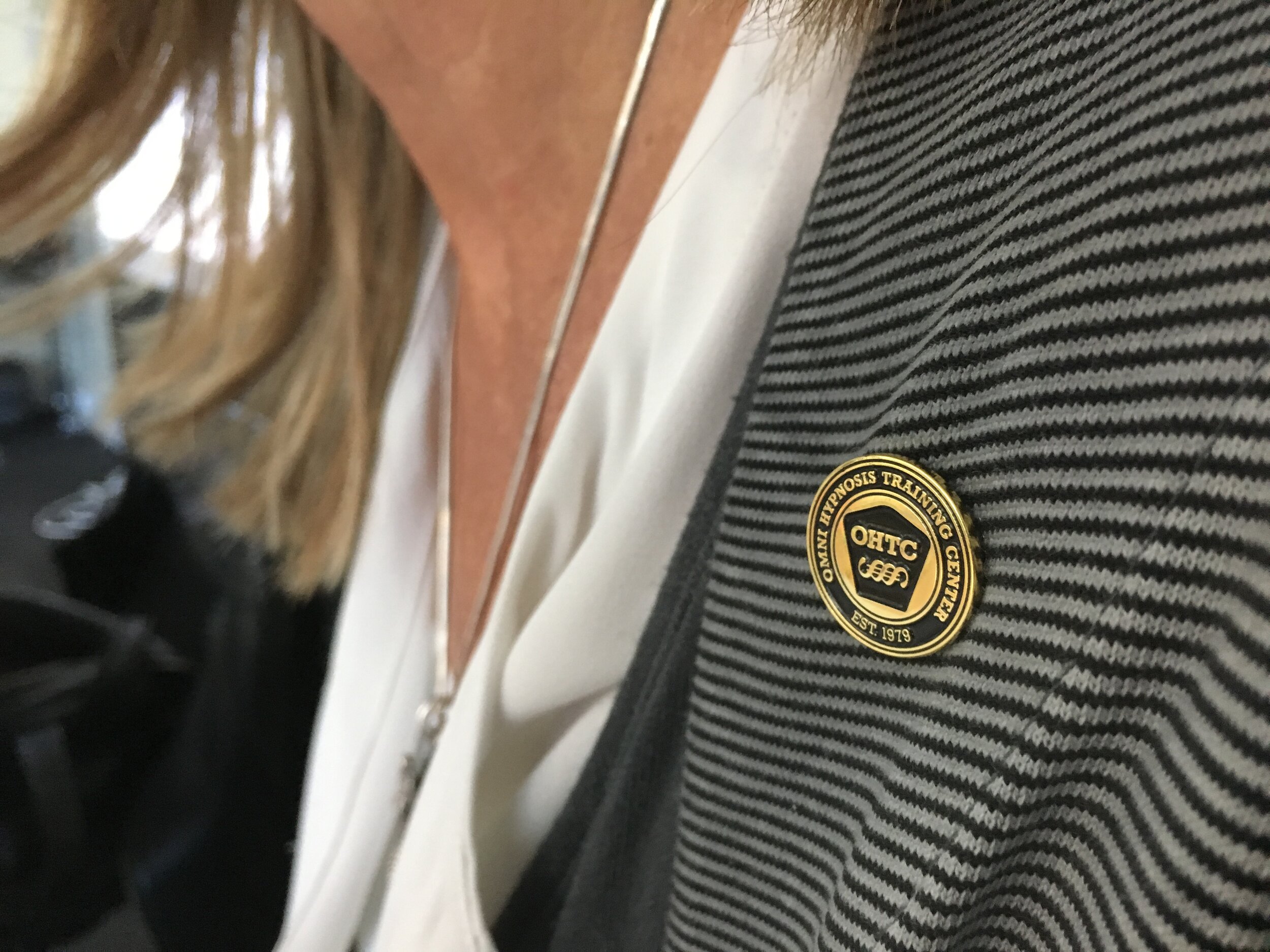 Un badge rond avec du texte en anglais et en français fixée sur un vêtement à rayures noires et grises. Le badge indique "OHTC" et mentionne un centre de formation pour les opérations de lyndon, créé en 1979.