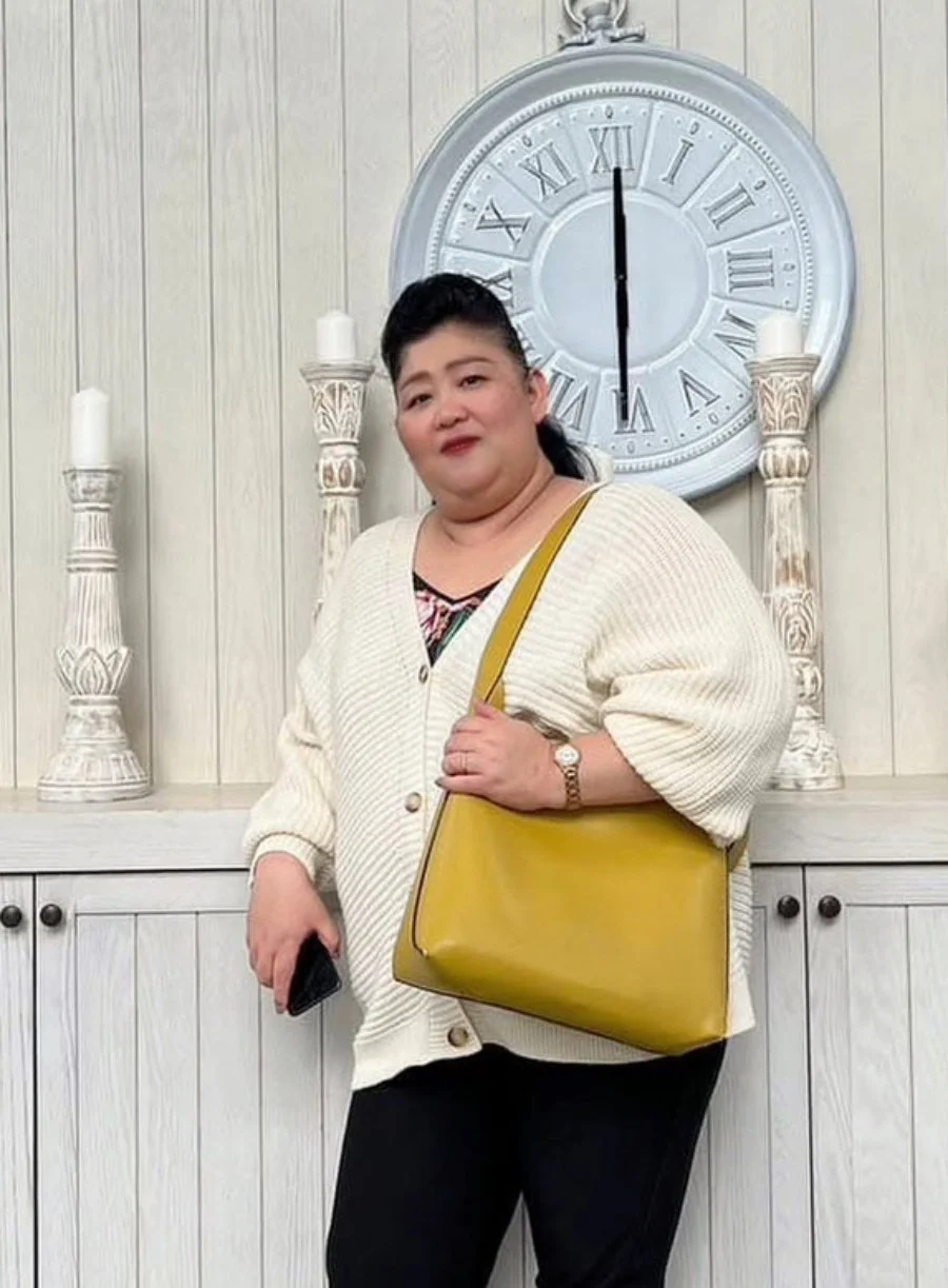 A woman standing in front of a large clock on a beige wall, holding a black phone, wearing a cream cardigan, black pants, a watch, with a yellow purse over her shoulder, and two candles on ornate pedestals on either side of her.
