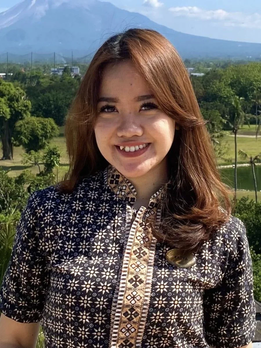 A woman with long, wavy, reddish-brown hair smiling outdoors, wearing a black and beige patterned traditional dress with a name tag, against a backdrop of lush green trees, a body of water, and mountains.