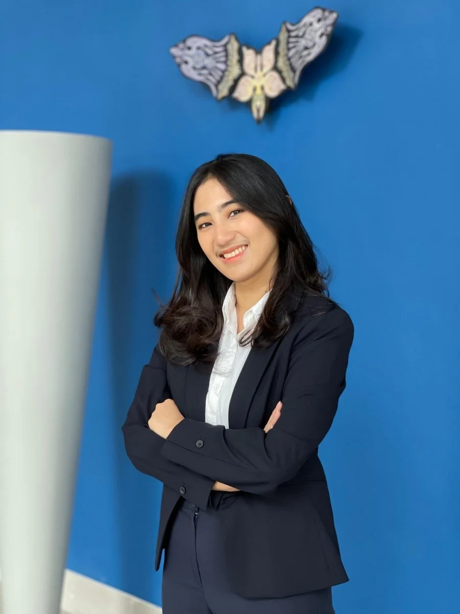 A woman in a business suit with crossed arms, smiling, standing against a blue wall with a decorative butterfly on it.