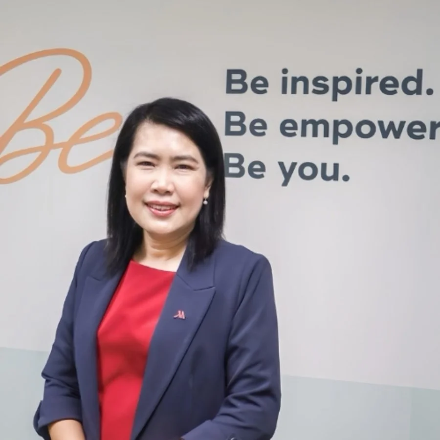 A woman with black hair smiling, wearing a navy blazer and red top, standing in front of a wall with inspirational quotes.