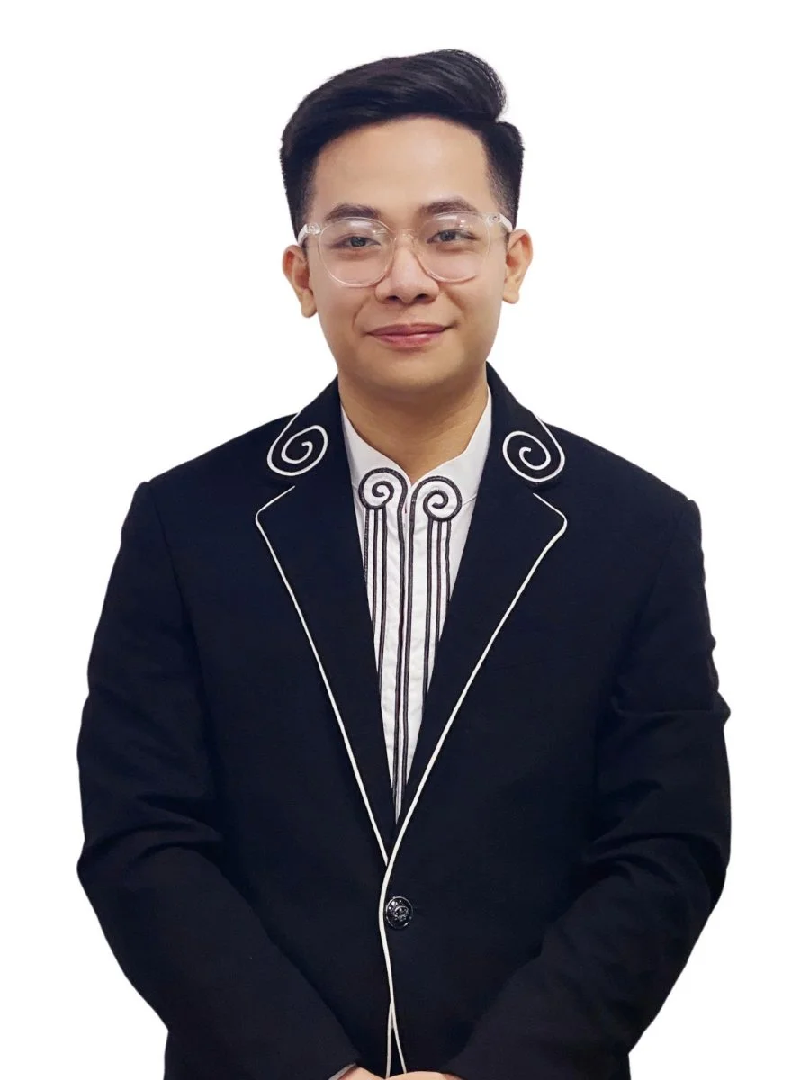 A young man in a black tuxedo with white piping and decorative embroidery, wearing glasses, stands in front of a white background, smiling softly at the camera.