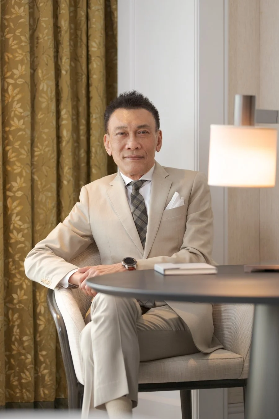 A middle-aged man in a beige suit, striped tie, and white shirt sitting at a round table in a well-lit room with gold patterned curtains behind him.
