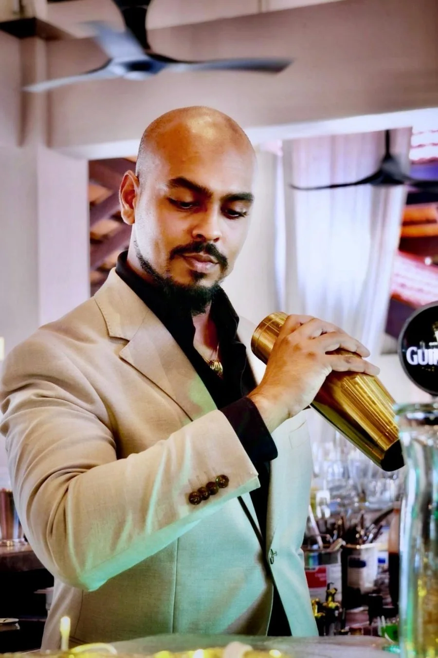 A man in a beige suit and black shirt holds a gold cocktail shaker in a bar or restaurant setting.