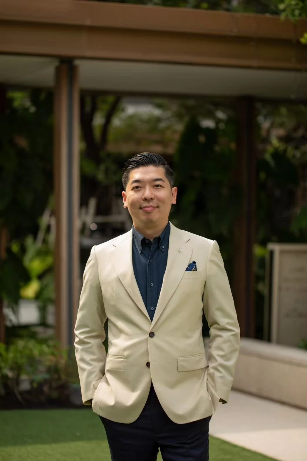 A man in a cream-colored blazer and dark shirt standing outdoors with greenery and modern architecture in the background.
