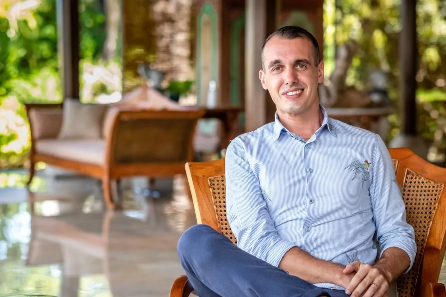 Young man smiling sitting on a wooden chair in a bright, outdoor lounge area with greenery in the background.