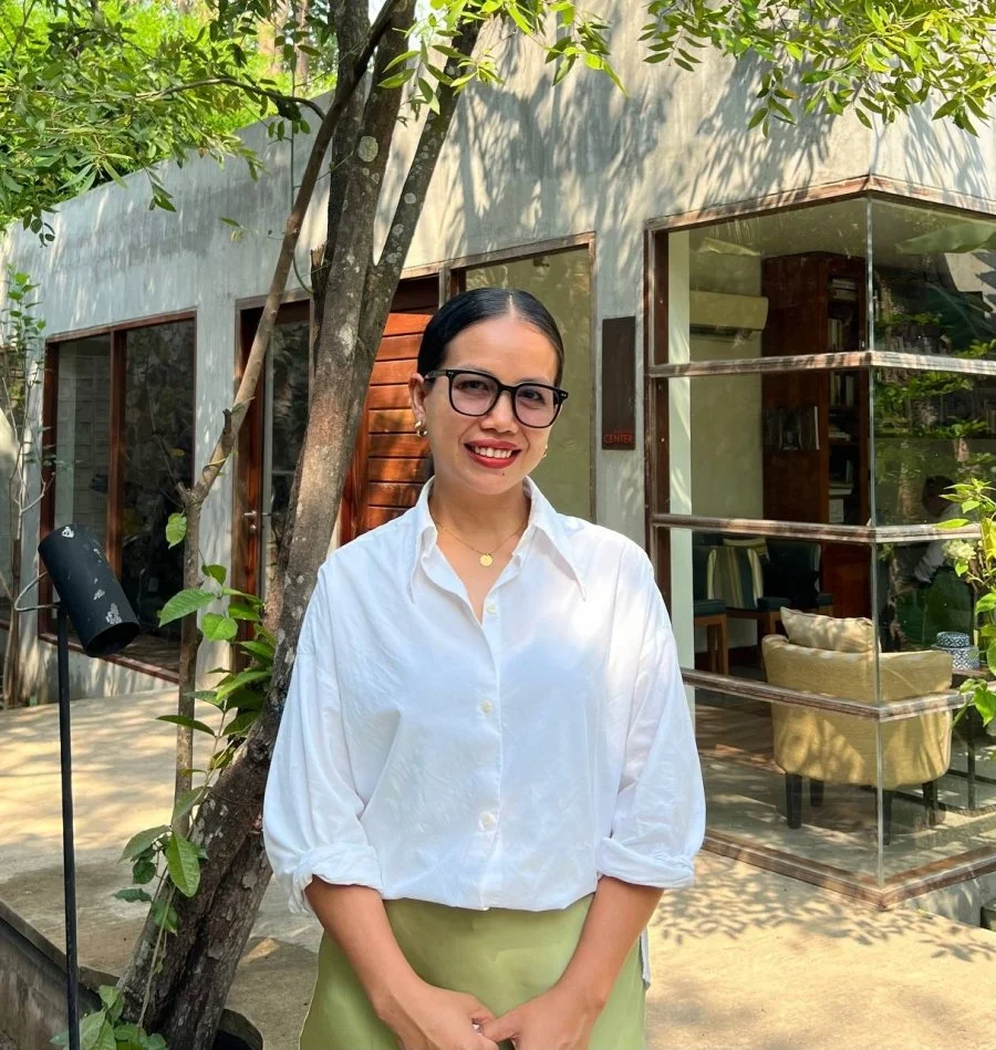 A woman with black hair, glasses, and earrings is standing outdoors in front of a modern building with glass windows and wooden accents, surrounded by trees and greenery.