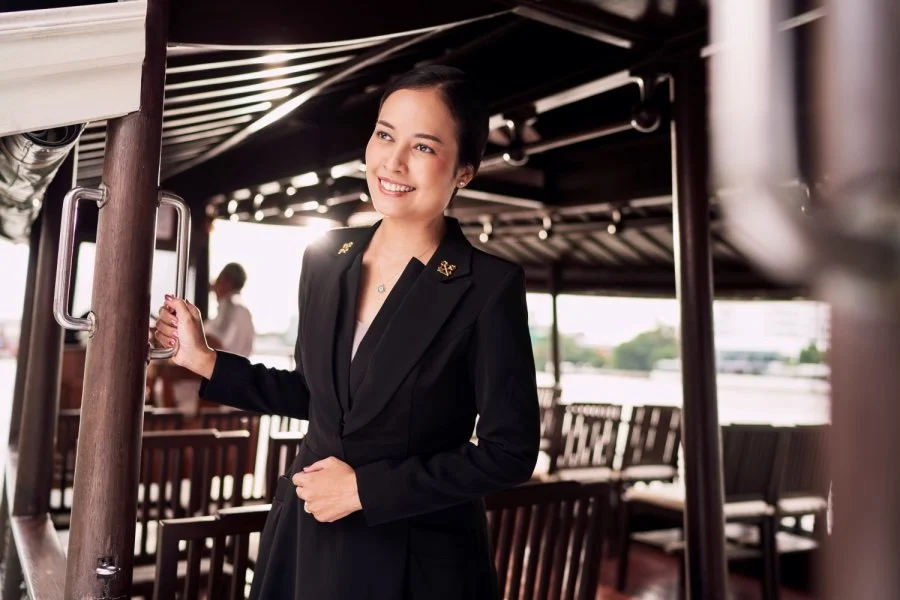 A smiling woman in a black blazer standing on a boat deck, holding onto a metal railing, with a waterfront view and other people in the background.