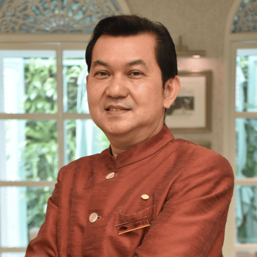 A man wearing traditional Asian attire posing indoors with a background of windows and greenery.