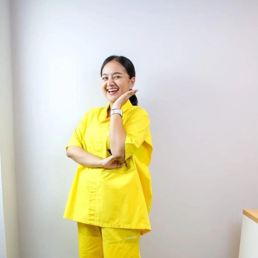 Young woman in yellow medical scrubs smiling and posing with hand on cheek in medical setting.