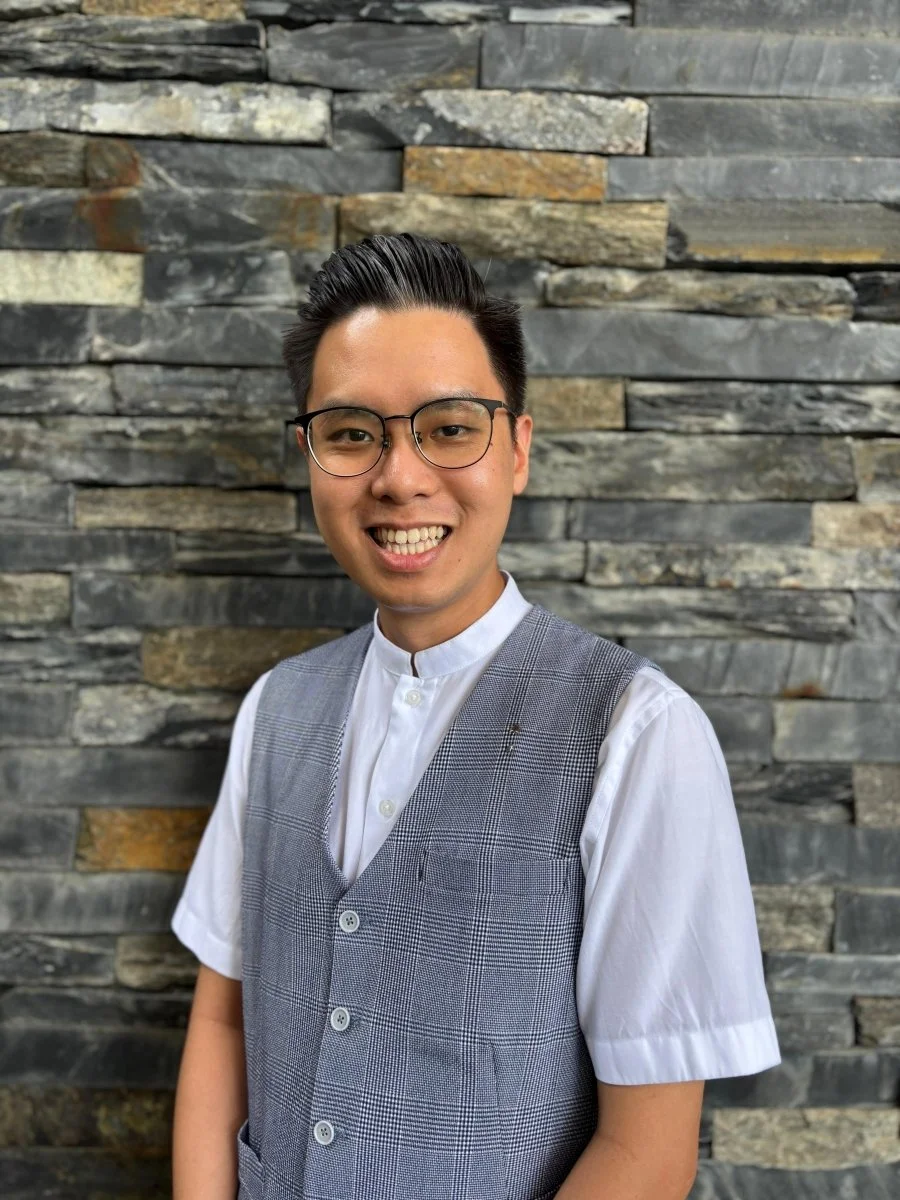 A young man with black hair, glasses, and a friendly smile, wearing a white shirt with a gray plaid vest, stands in front of a textured stone wall.