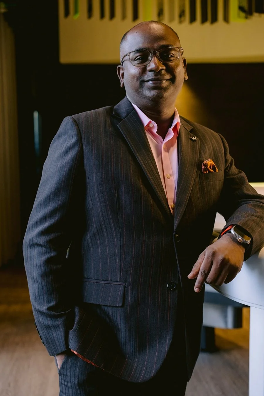 A man in a dark pinstripe suit, pink shirt, and glasses standing indoors with one hand on a white counter, smiling and looking at the camera.